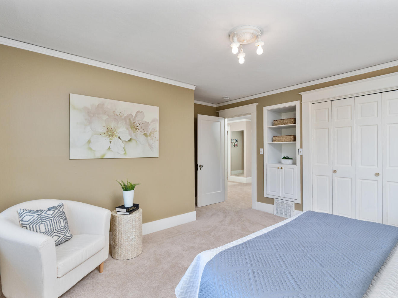 A cozy bedroom with beige walls, a bed with a blue quilt, a white chair with a patterned cushion, and a round woven side table. Theres a floral painting on the wall and built-in shelving next to a closet with white sliding doors.