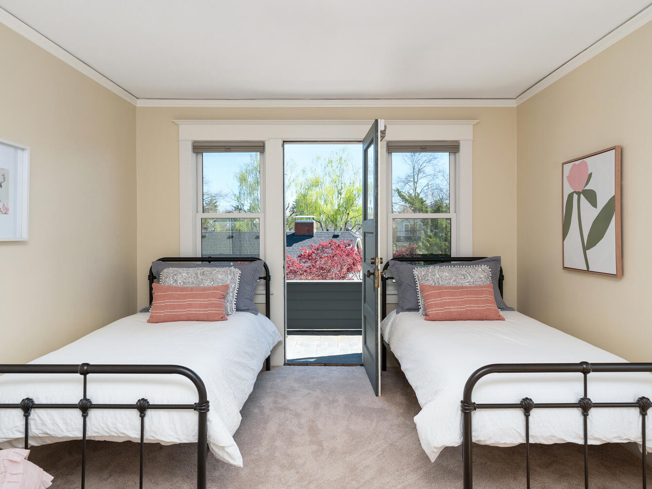 A bedroom with two twin metal beds, white bedding, and pink accent pillows. A door in the middle of the room opens to a view of a patio with greenery and flowers. The walls are beige with framed flower artwork.