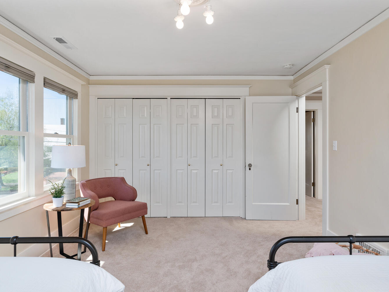 A bright bedroom featuring a comfortable pink chair with a small side table and book. Theres a large closet with white paneled doors, neutral walls, and carpeted flooring. Two partial beds are visible at the bottom corners. Natural light streams in through the windows.