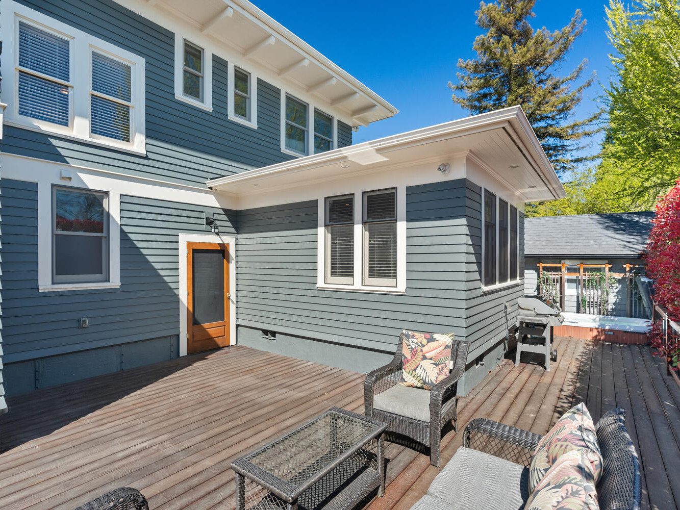 A spacious wooden deck with wicker furniture and floral cushions is attached to a two-story gray house. White-trimmed windows line the exterior walls. Lush greenery and a bright blue sky are visible in the background.