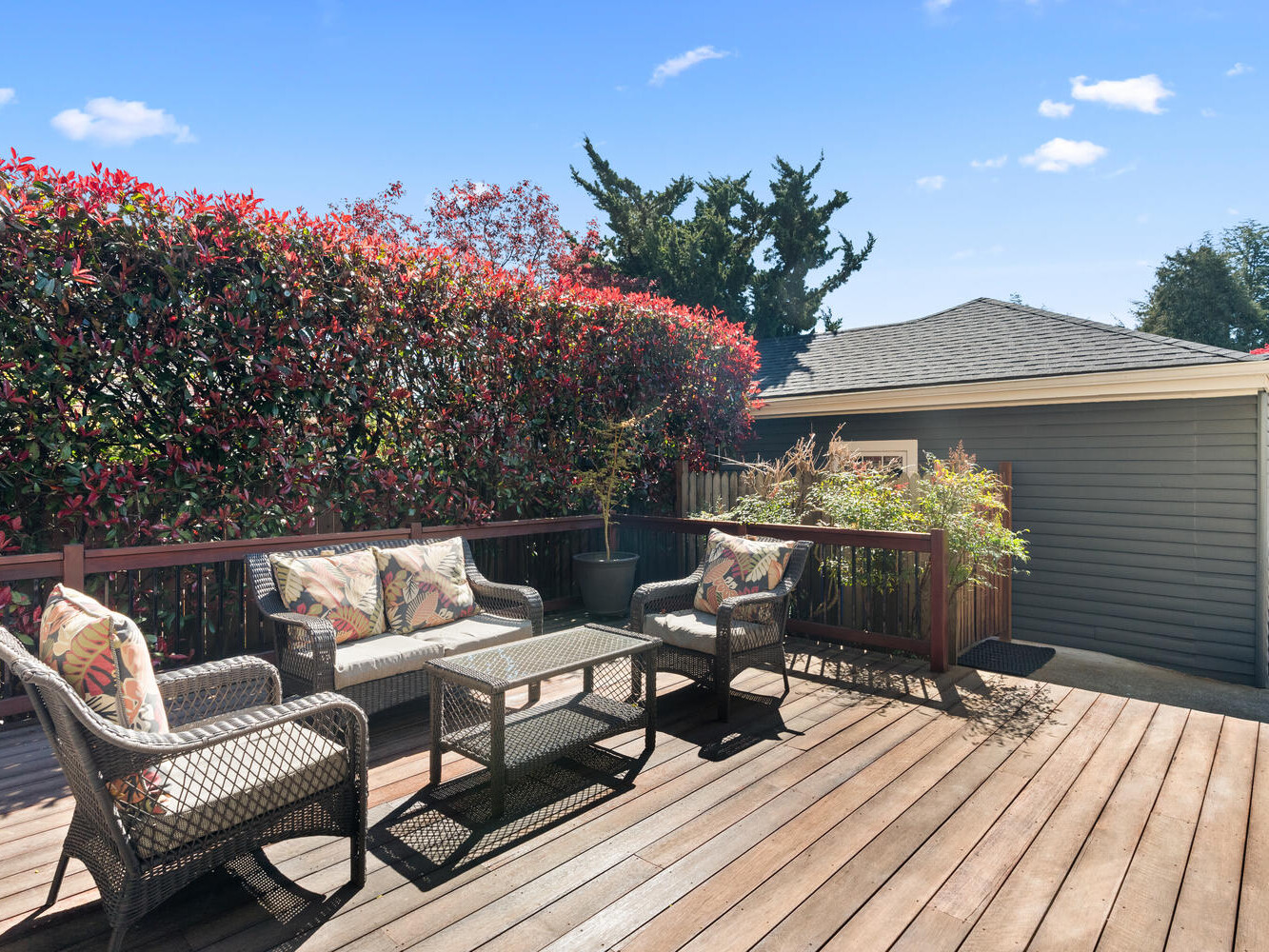 A sunny wooden deck with wicker furniture, including a sofa, two chairs, and a glass-top coffee table. The deck is bordered by lush red and green bushes, and a gray wooden house is visible in the background. Clear blue sky above.