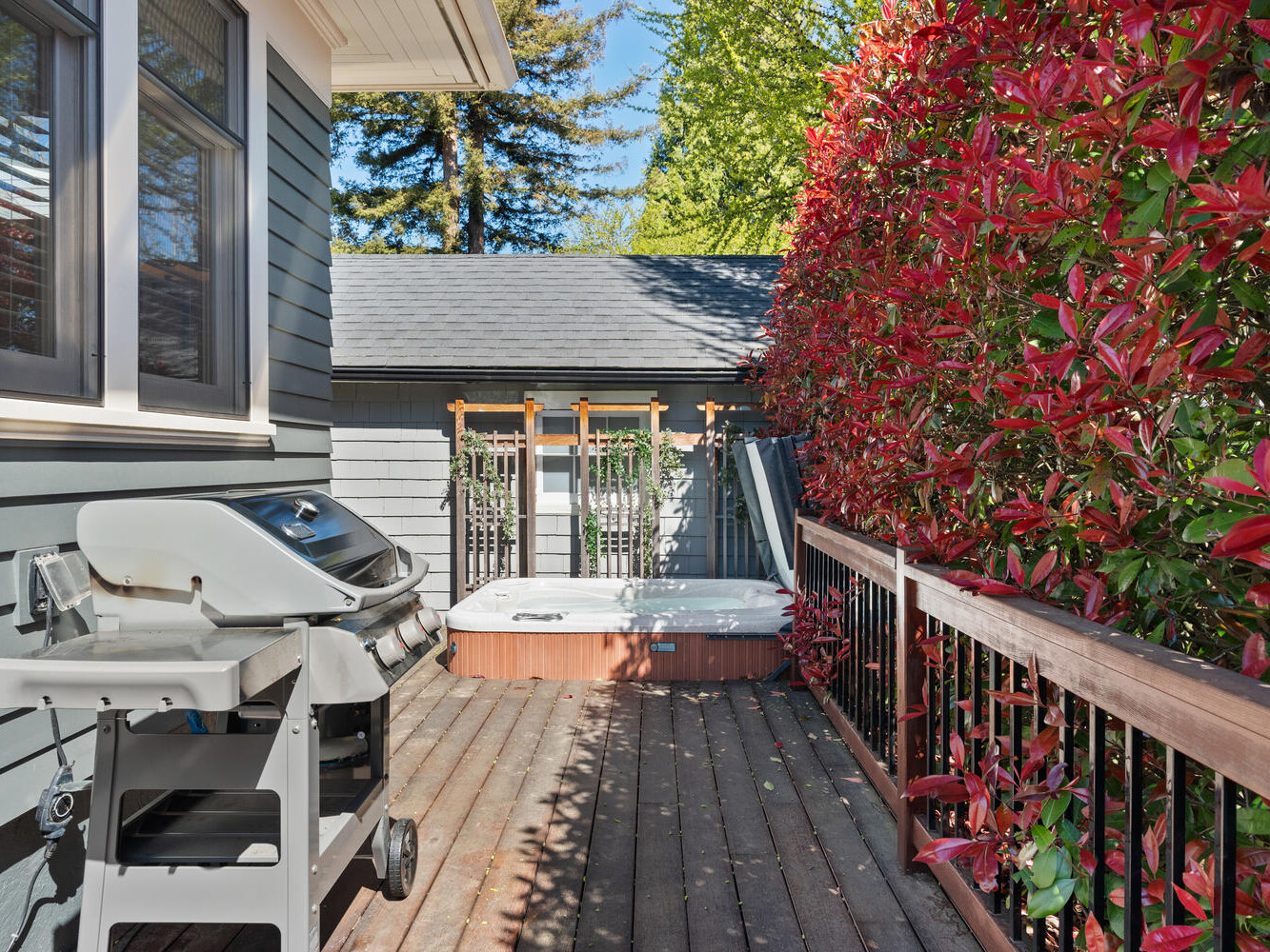 A wooden deck with a barbecue grill and a hot tub. The deck is bordered by lush red-leaved bushes, and a building with gray siding is in the background under a clear blue sky.