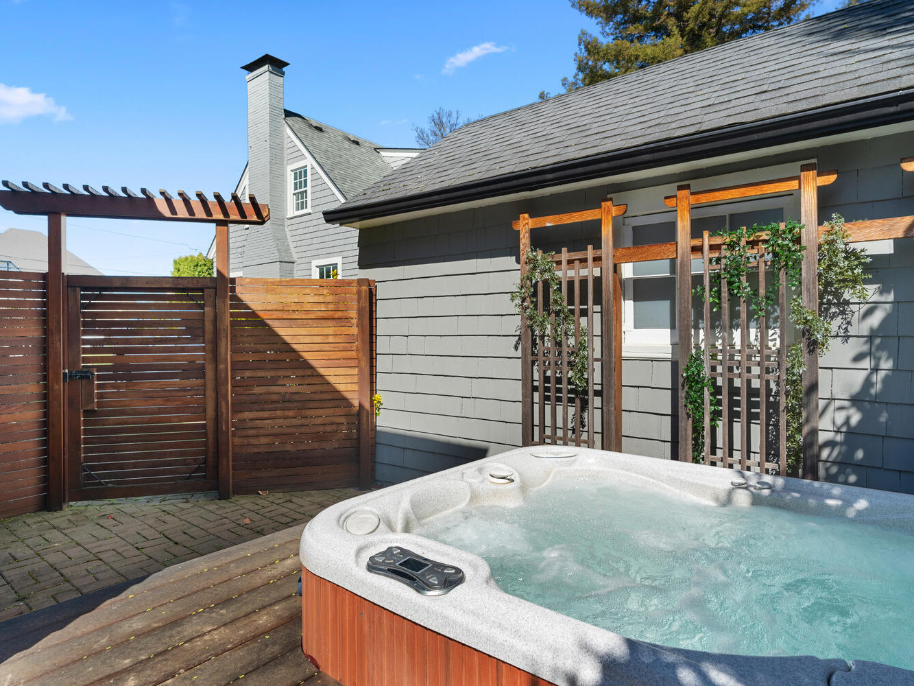 A backyard scene featuring a hot tub with bubbling water. The area is enclosed by a wooden privacy fence and has a small trellis with plants. The weather is sunny with a clear blue sky.
