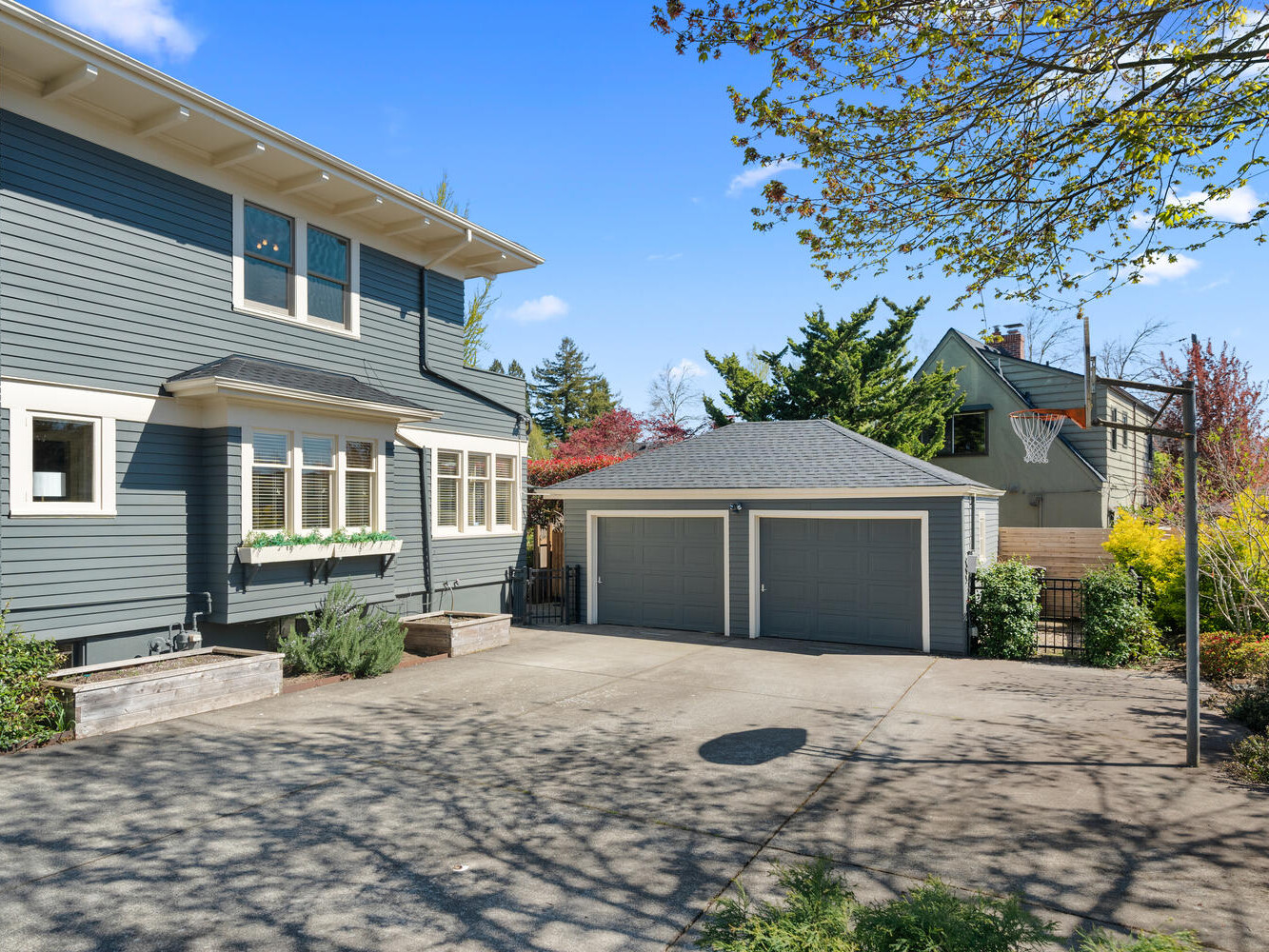 A two-story house with gray siding and white trim features a double garage. The driveway is paved, and a basketball hoop is installed near the garage. Surrounding trees and a clear blue sky add to the suburban setting.