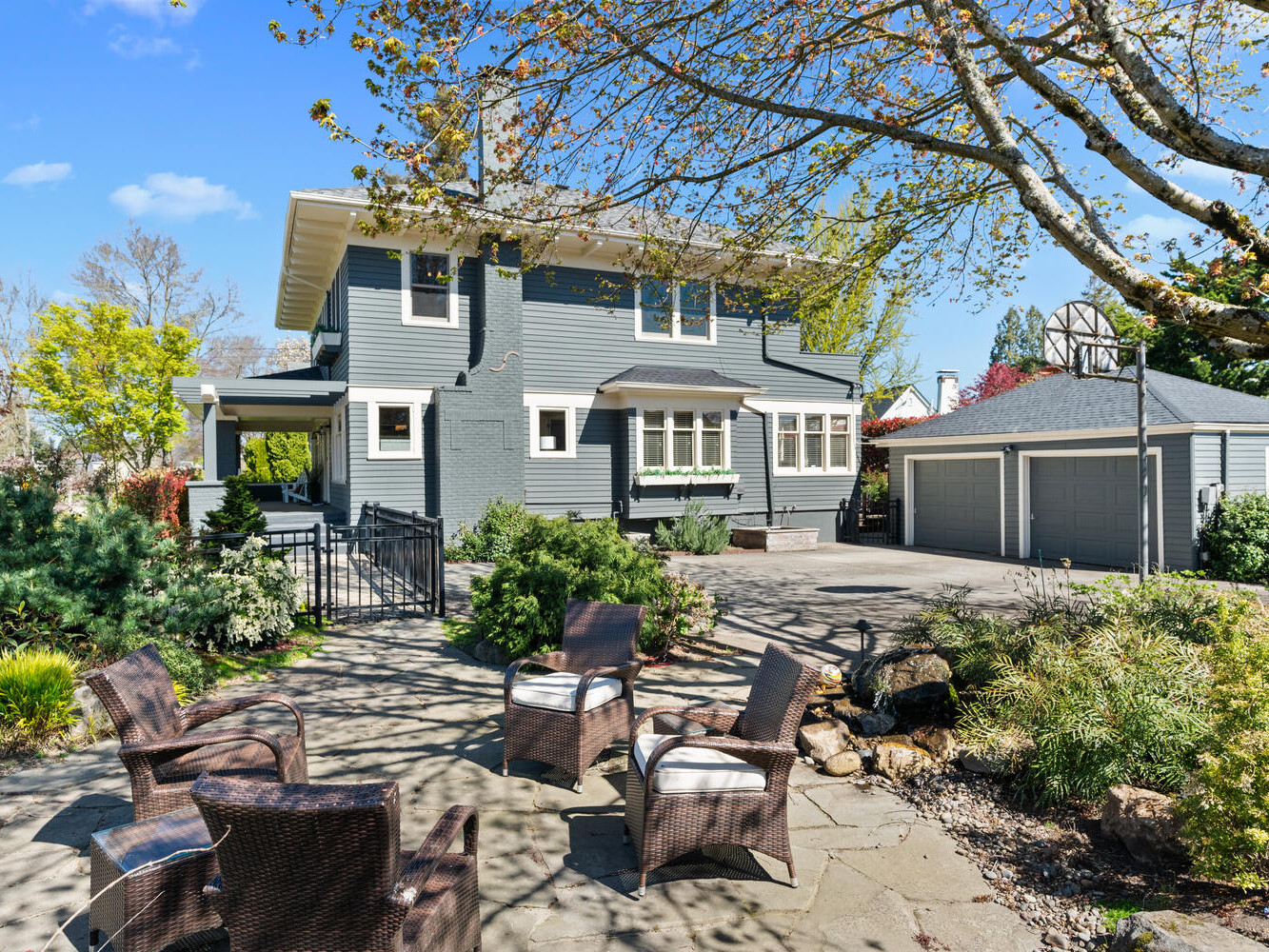 A two-story gray house with white trim is surrounded by a lush garden. In the foreground, there’s a patio with wicker chairs. A detached gray garage is visible to the right, and the sky is clear and blue.