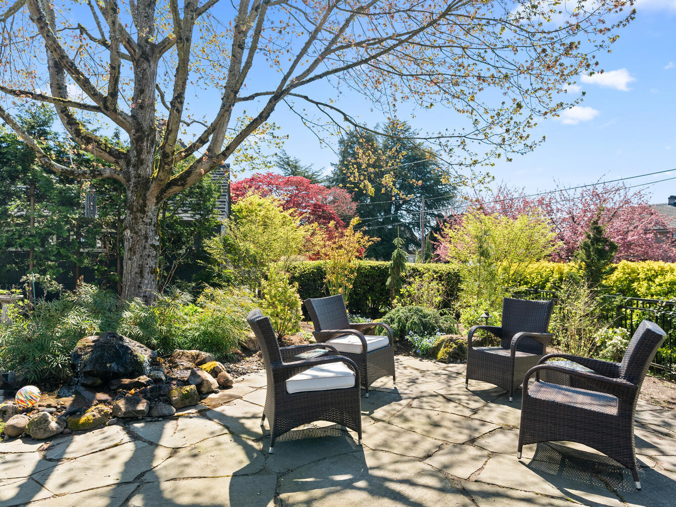 A serene patio setting featuring four wicker chairs arranged around a small water feature with rocks. Surrounded by lush greenery, a large tree provides shade, and the background showcases colorful shrubs and a clear blue sky.