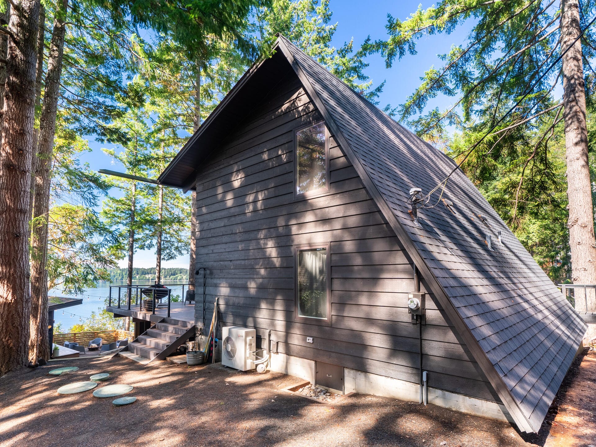 A dark wooden A-frame cabin sits among tall trees with a view of a lake and the sky in the background. The cabin has a sloped roof and several windows. A wooden deck extends from the side, surrounded by forest scenery.