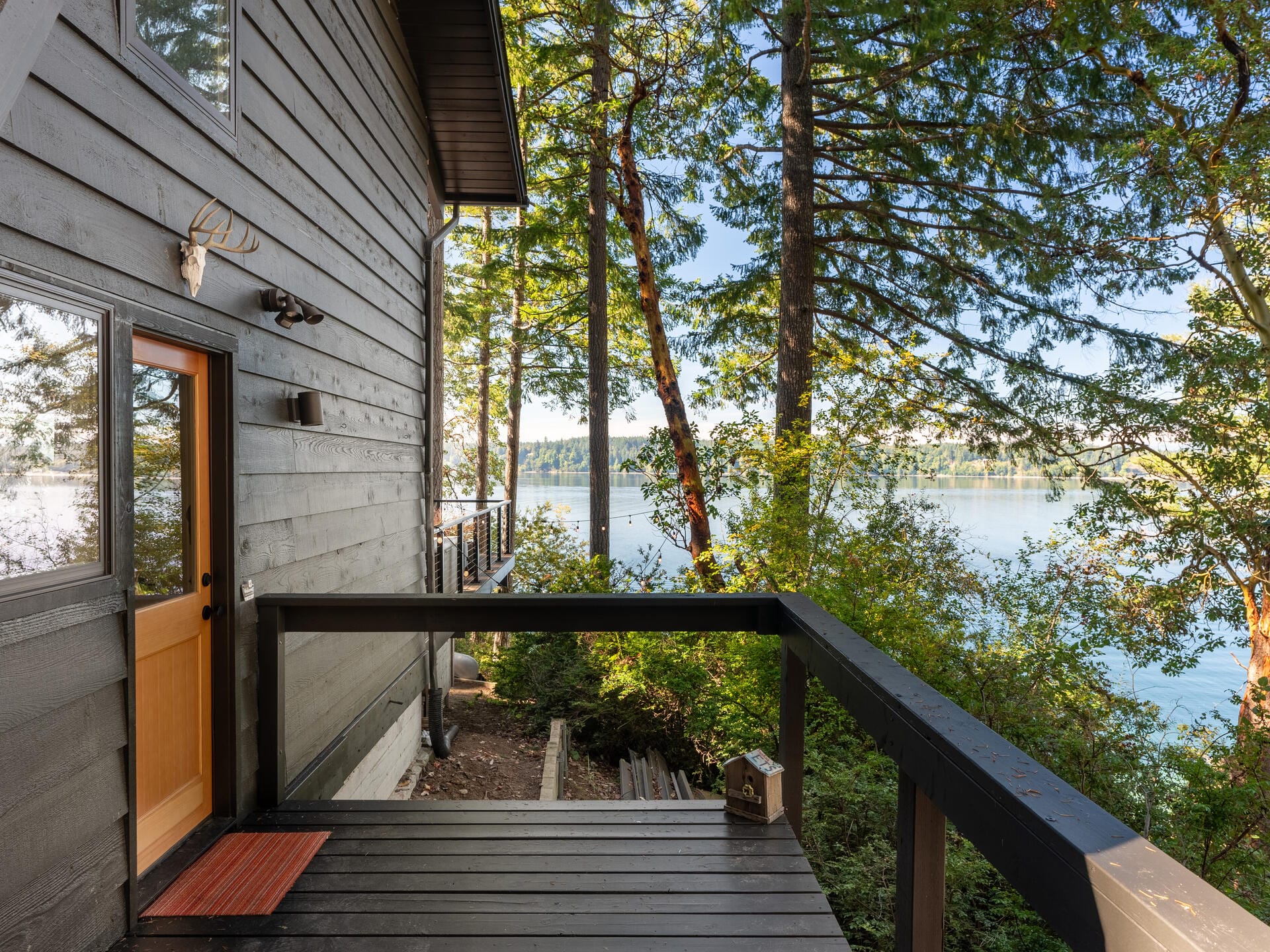 Wooden cabin with a balcony overlooking a serene lake, surrounded by tall trees. A door with a small window and antler decor is visible. Bright, sunny day with calm water in the distance.