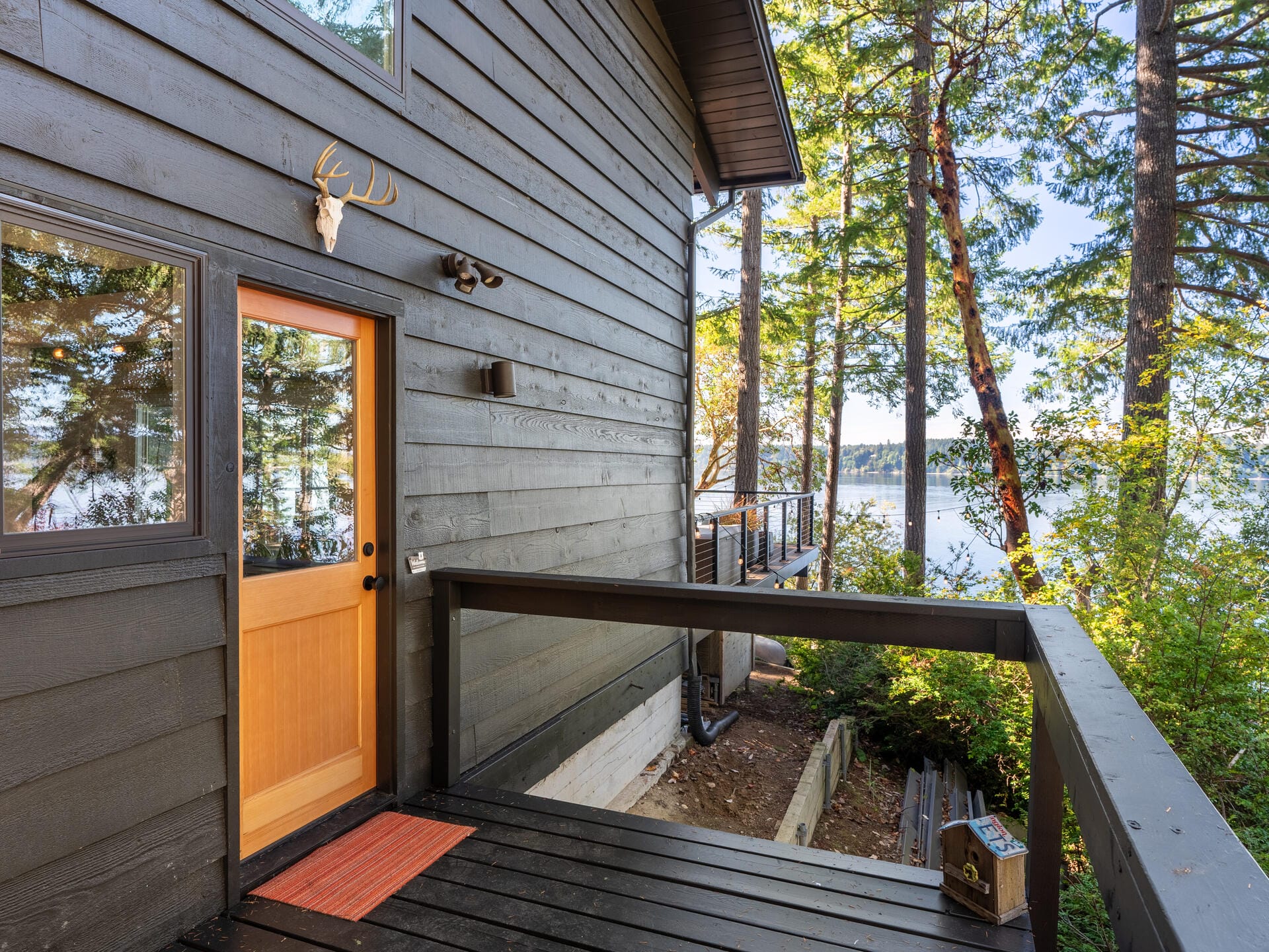 A cozy wooden cabin with a natural wood door and a deer antler decoration above it. The cabin is surrounded by tall trees and overlooks a scenic lake, visible through the forest. A pathway leads to the entrance, and the scene is peaceful and inviting.