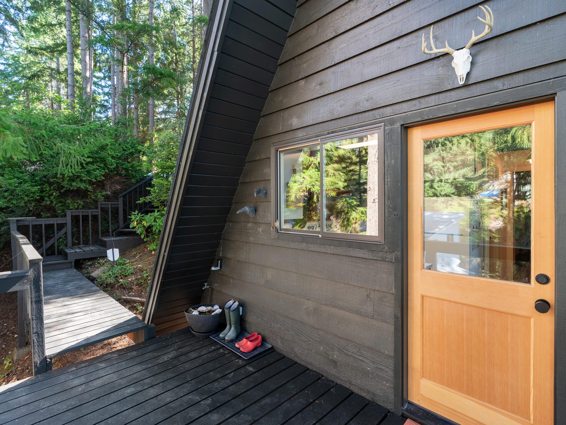 A black A-frame cabin with a wooden door and a deer skull decoration above it. Red and black boots are on the dark wooden porch, and a curved walkway leads away, surrounded by lush green trees and foliage.