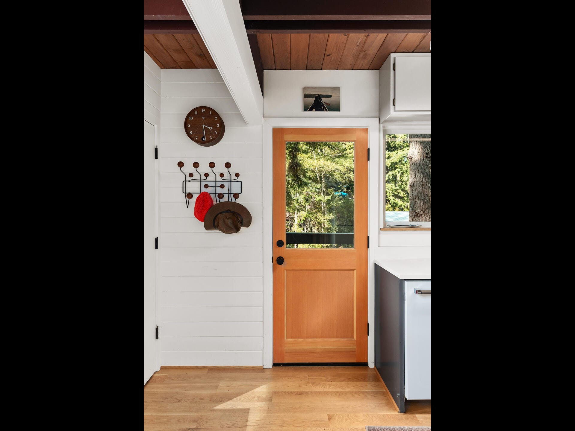 A cozy entryway features a wooden door with a glass pane, allowing a view of lush greenery outside. A wall-mounted rack holds coats and hats, and a round clock hangs nearby. The interior has white panel walls and a wooden floor.