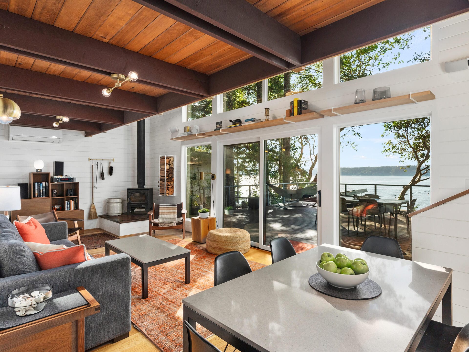 A modern living room with large windows showing a lake view. The space features a gray couch, wooden ceiling beams, a dining table with green apples, and a sliding door leading to a deck with outdoor furniture.
