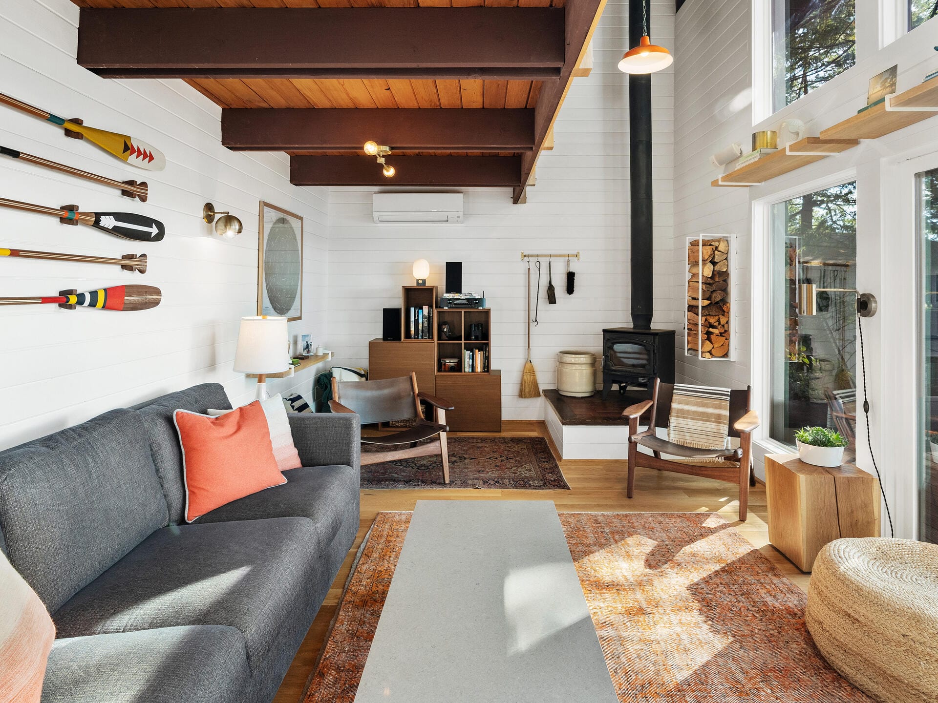 A cozy living room with a gray sofa adorned with orange and white pillows. Wooden beams and a high ceiling add warmth. Decorative paddles on the wall, a wood-burning stove, and large windows create a rustic atmosphere. A patterned rug covers the floor.