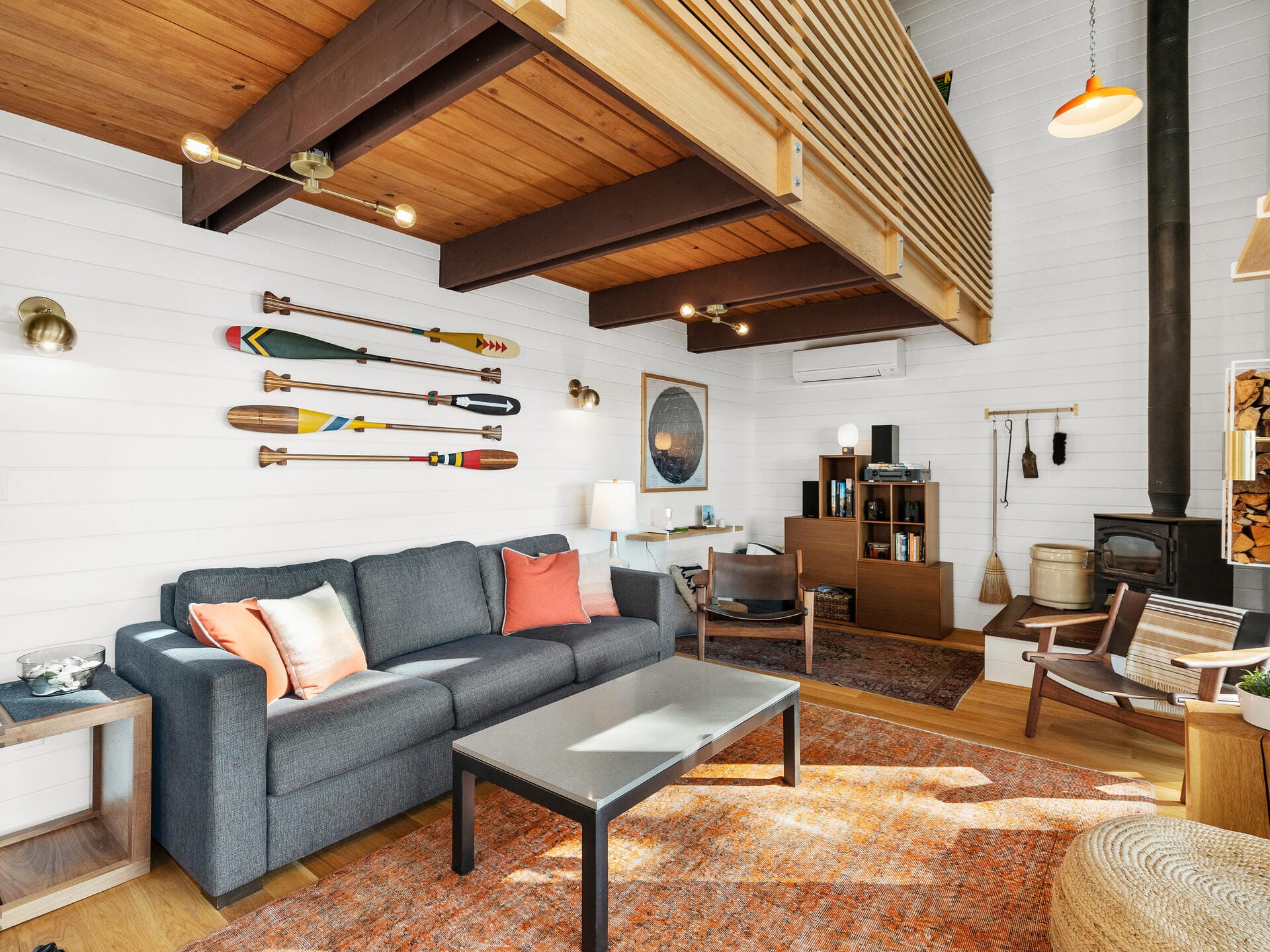 A cozy living room with a gray sofa and orange cushions, a modern coffee table, and colorful oars mounted on a white shiplap wall. A wood-burning stove and warm wood accents complement the space, with natural light streaming in.