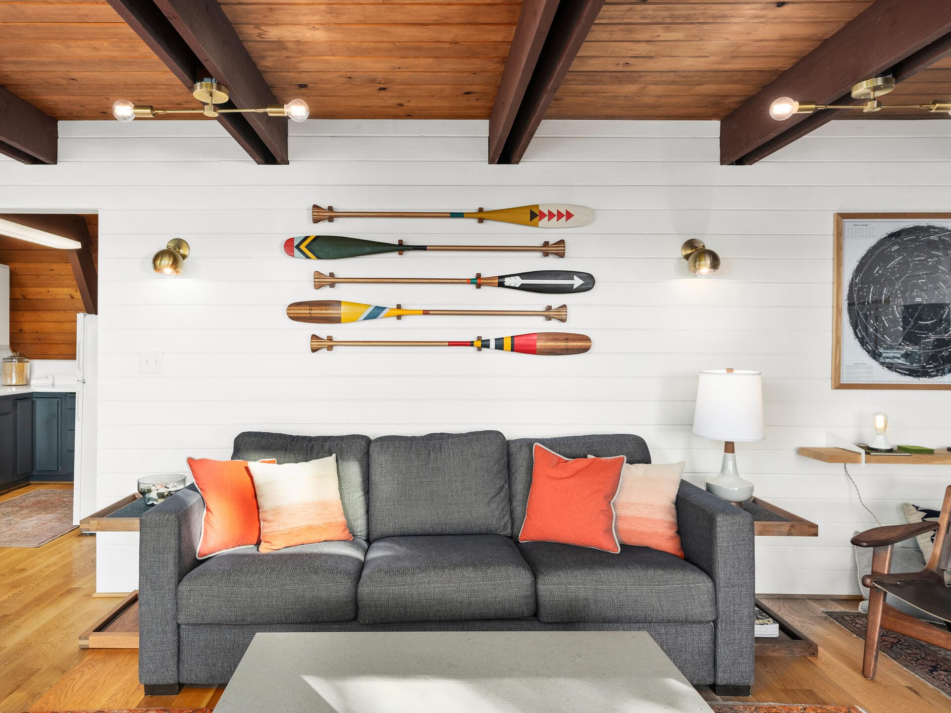 A cozy living room with a gray sofa adorned with orange and beige pillows. Four decorative paddles hang on a white shiplap wall above the sofa. The wooden ceiling features exposed beams, and there are modern wall sconces beside the paddles.