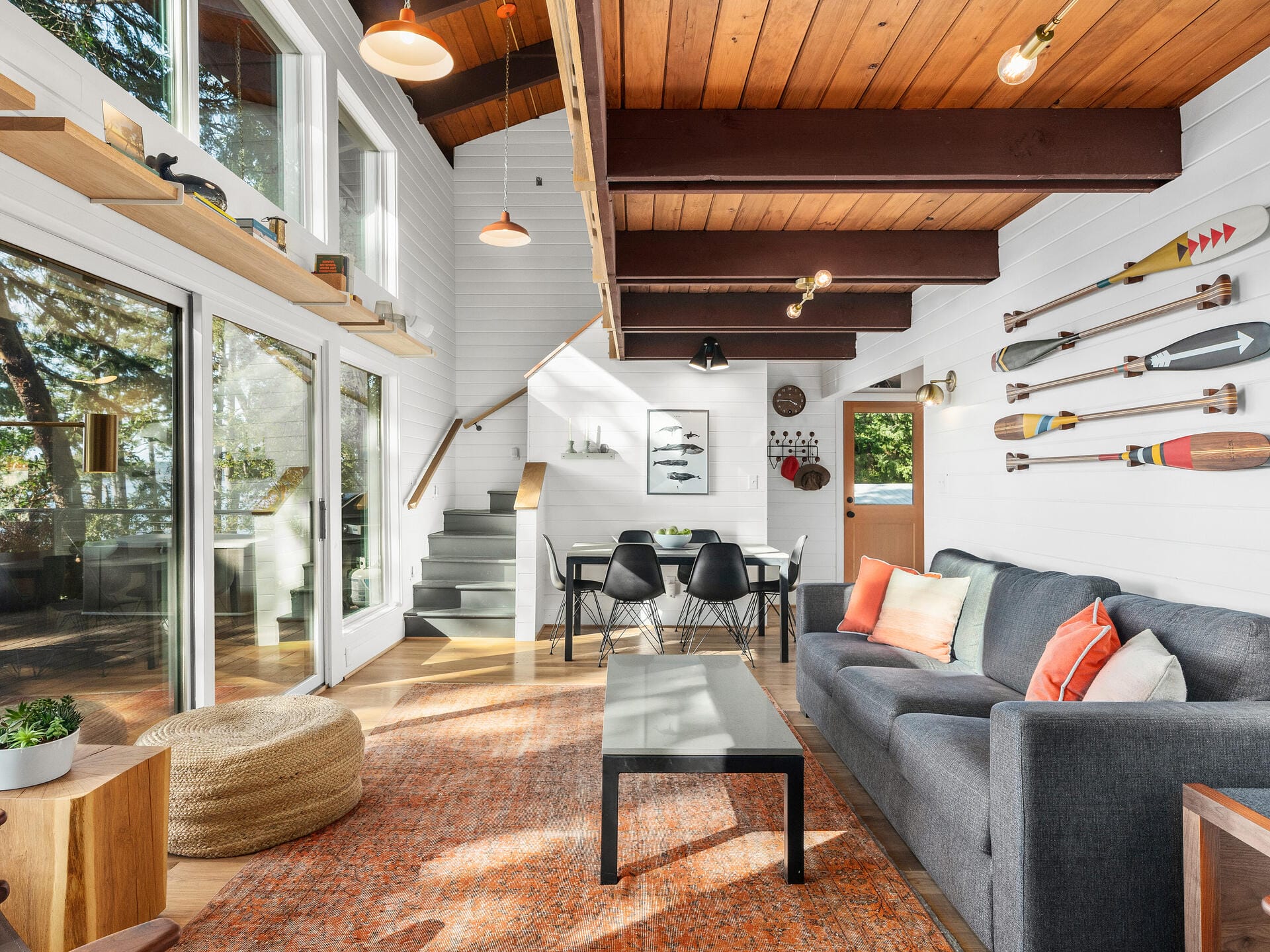 A modern living room with a high ceiling features a gray sofa, orange rug, and a wooden coffee table. Paddle decorations adorn the white walls. Large windows let in natural light, and a dining table with black chairs is in the background.