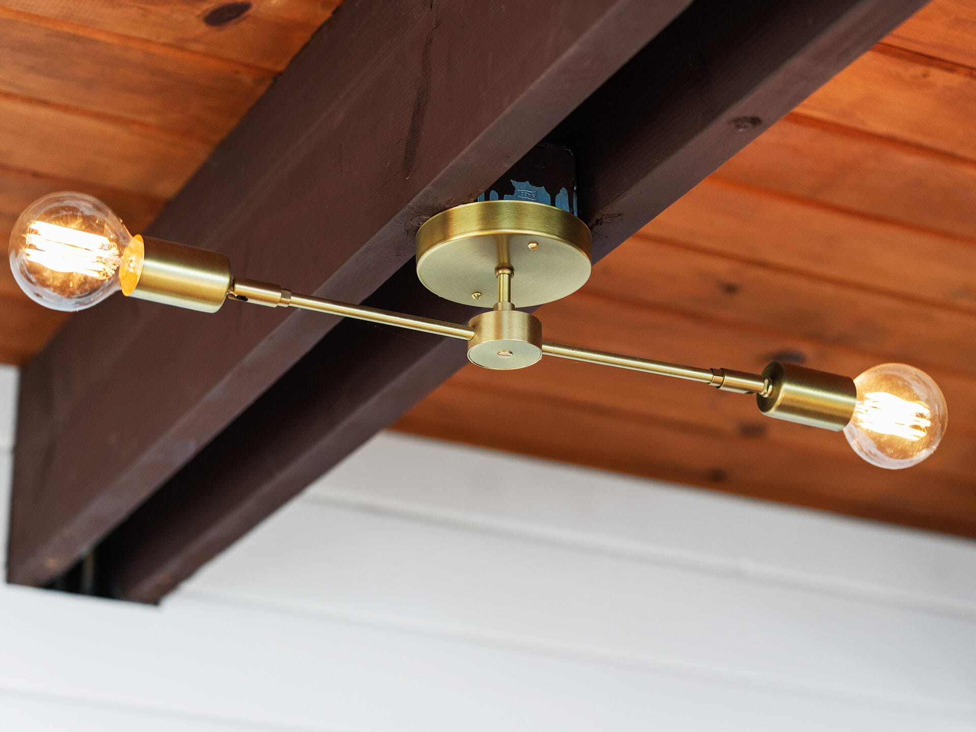 A gold, two-bulb ceiling light fixture with exposed filament bulbs is mounted on a wooden ceiling beam. The wooden planks and white wall give the space a warm and rustic appearance.