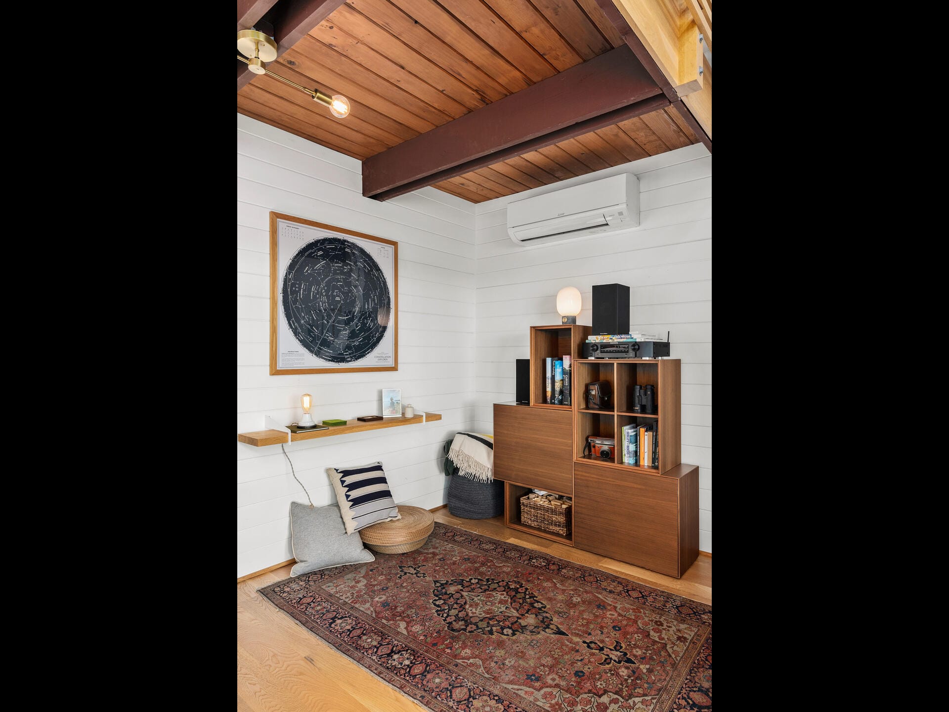 Cozy room with a mid-century vibe, featuring a wooden bookshelf with various decorative items, a patterned rug, a large abstract art piece on the wall, and a small cushioned seating area. The walls are white with wooden accents.