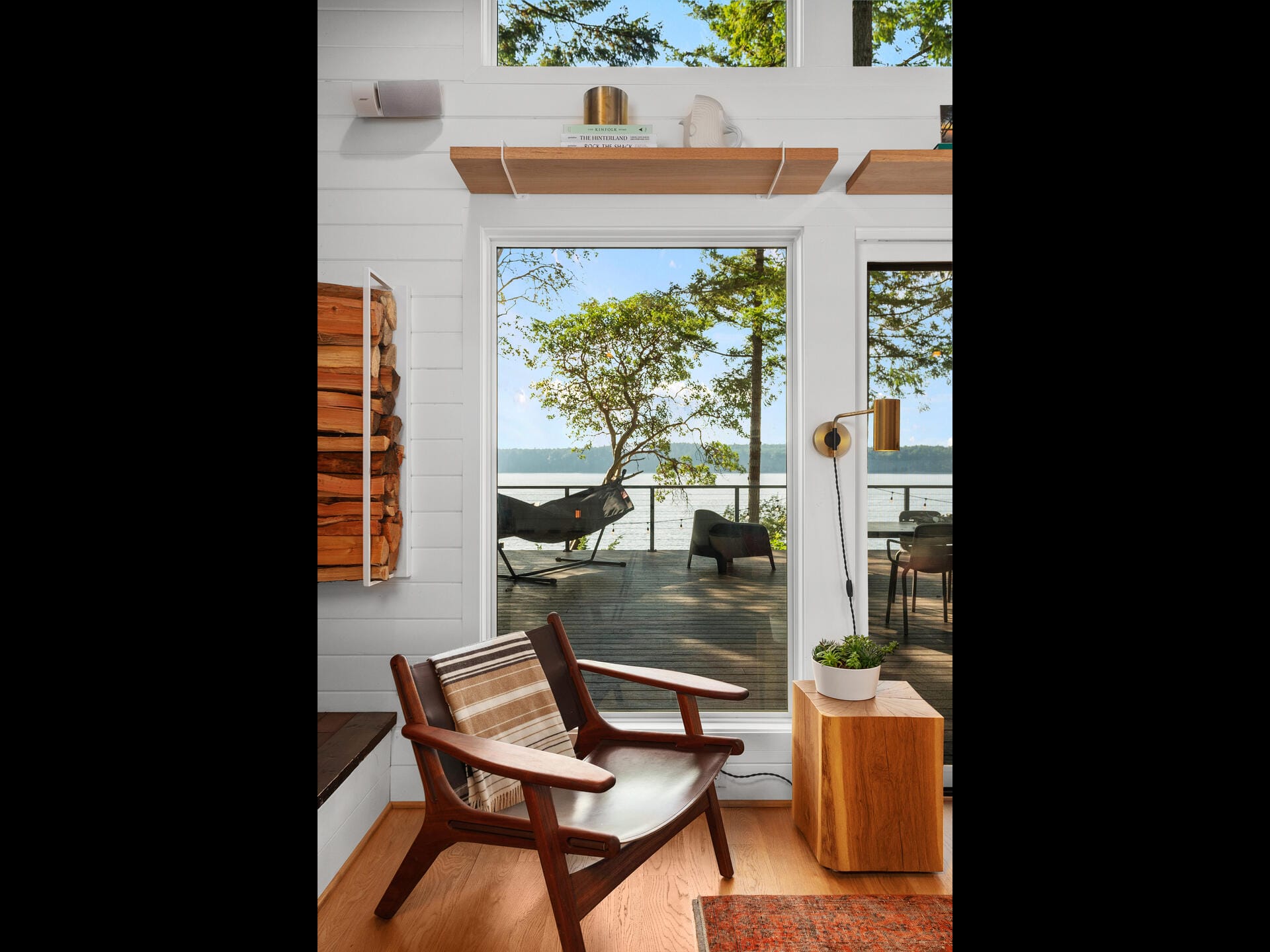 A cozy room with a modern wooden chair and a side table with a potted plant. Large windows offer a view of a deck with lounge chairs, trees, and a serene lake. Sunlight filters through, creating a warm and inviting atmosphere.