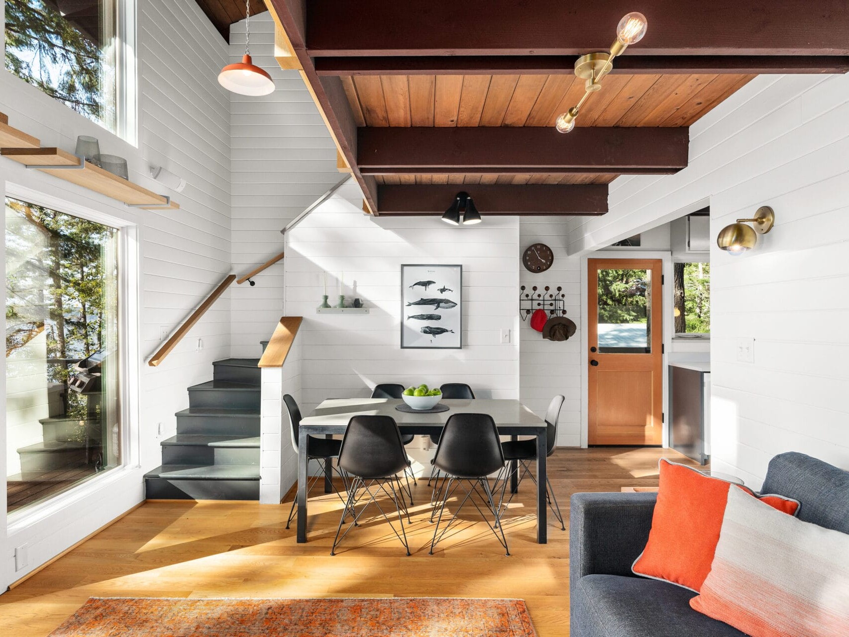 A modern living and dining area in a cozy Portland, Oregon home. Features include wooden beams, a staircase, large windows with a forest view, and a dining table with black chairs. Colorful cushions adorn the gray sofa, complemented by various wall art and decor pieces.