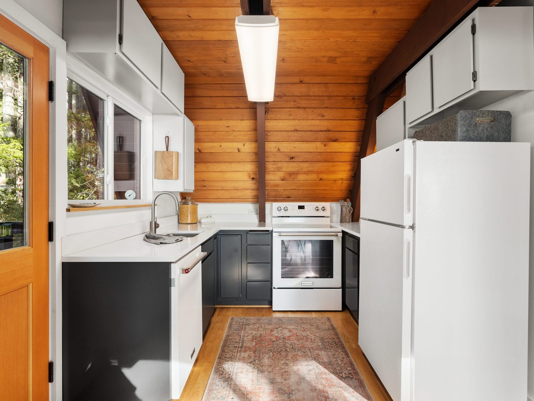 A cozy kitchen in Portland, Oregon, boasts a sloped wooden ceiling and white cabinets. It features a white fridge, oven, dishwasher, and sink. Natural light pours in through the door and window, while a patterned rug warms the wooden floor.