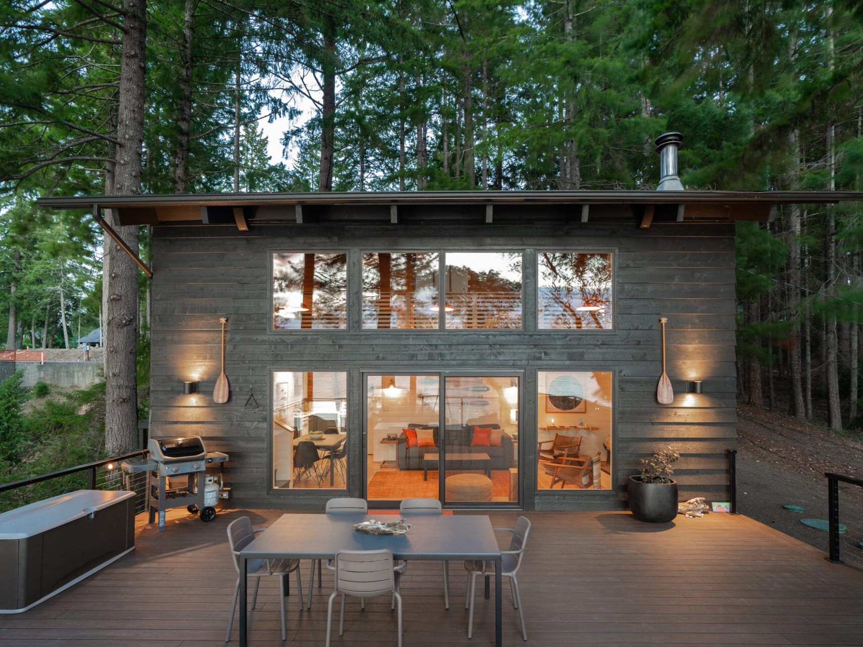A modern cabin with large windows is nestled in a forest setting near Portland, Oregon. The outdoor deck features a dining table with six chairs. The warm interior lighting and dense trees create a cozy and inviting atmosphere.