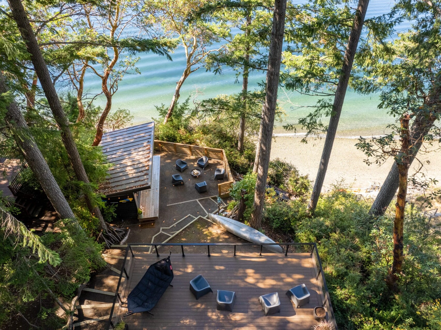 Aerial view of a serene beachside area reminiscent of Portland, Oregon, with tall trees, a wooden deck, and lounge chairs. There's a small cabin, a kayak nearby, and a sandy shore leading to calm, turquoise water.
