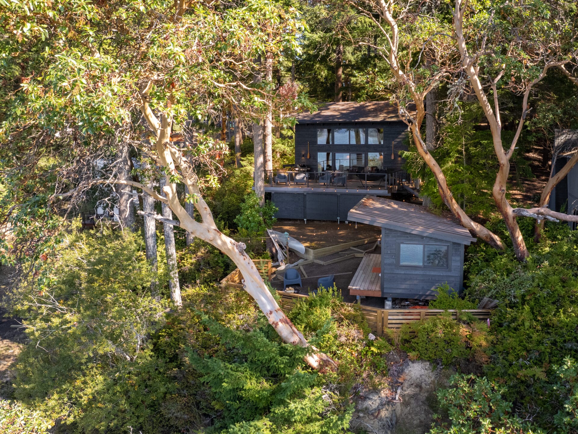 A modern black cabin with large windows is nestled among dense greenery and tall trees near Portland, Oregon. The multi-level structure features a wooden deck surrounded by lush foliage, perfectly capturing the serene forest setting unique to the Pacific Northwest.