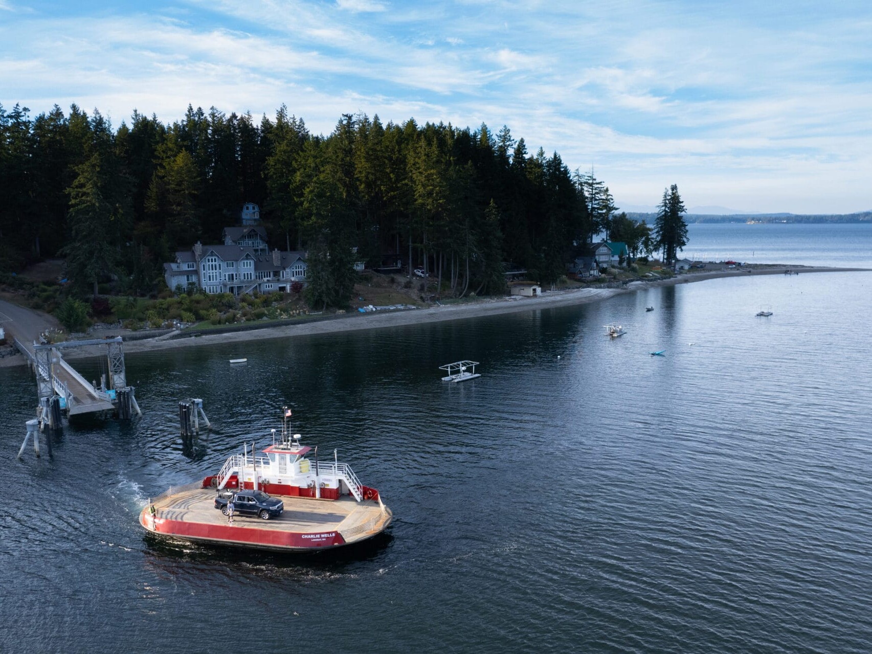 A car ferry with a red and white design approaches a wooded shoreline reminiscent of Portland, Oregon, dotted with several houses. The scene captures calm waters under a partly cloudy sky, with a dock extending into the water. Nearby, a small seaplane floats peacefully.
