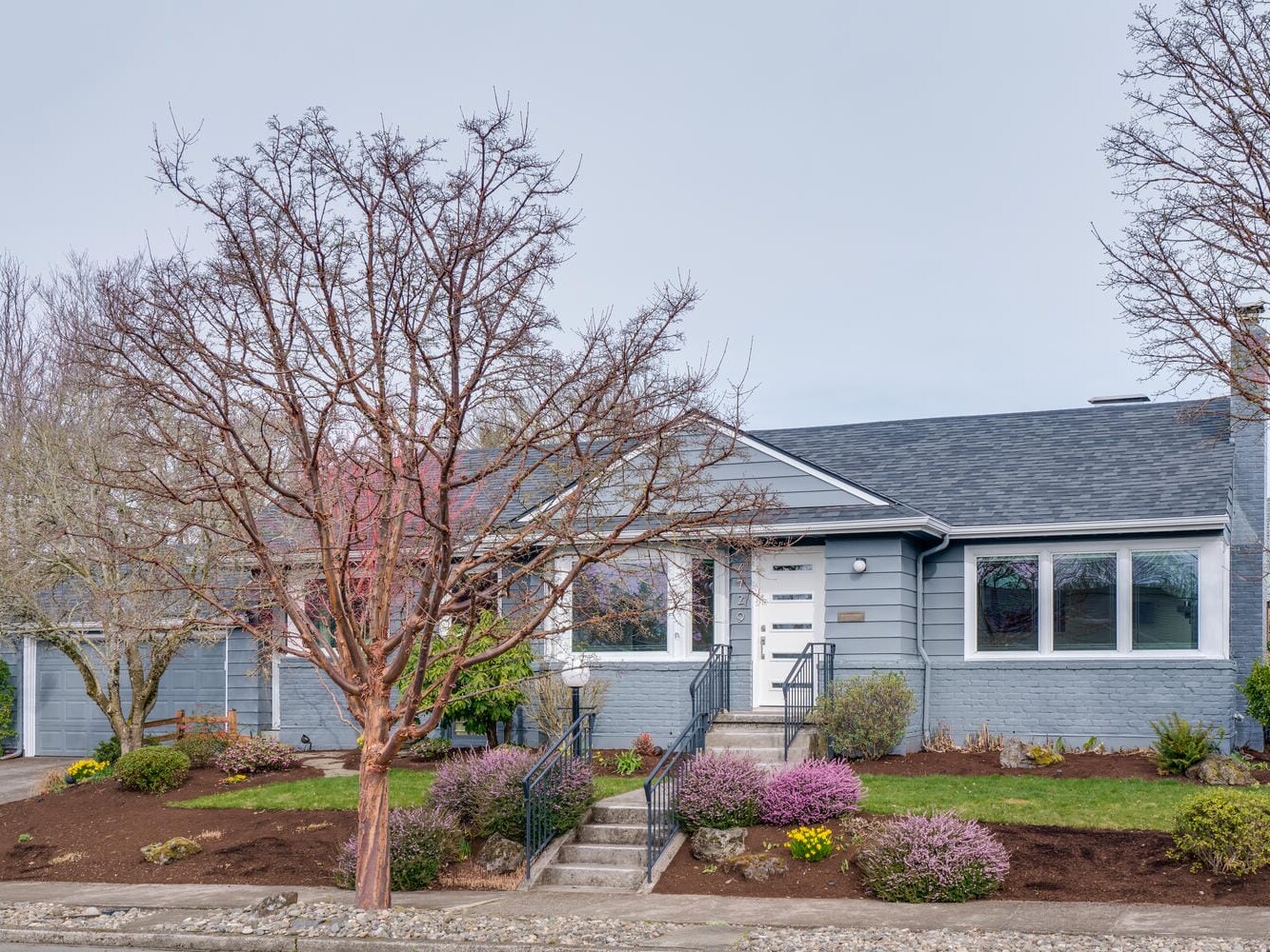 A blue-gray single-story house with a dark gray roof sits serenely in Portland, Oregon, surrounded by a landscaped garden. Leafless trees and flowering bushes adorn the front yard, with a paved walkway guiding visitors to the entrance. The overcast sky adds a touch of Pacific Northwest charm.