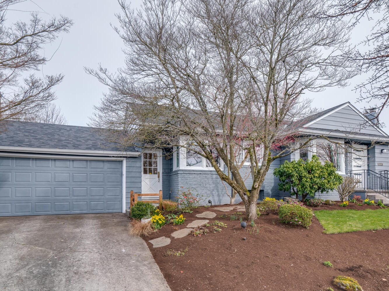 A blue-gray suburban house in Portland, Oregon, boasts a two-car garage and sits amidst bare trees with a garden of bushes and plants. A stone path leads to the front entrance, with a yard partially mulched and featuring a lush green lawn.