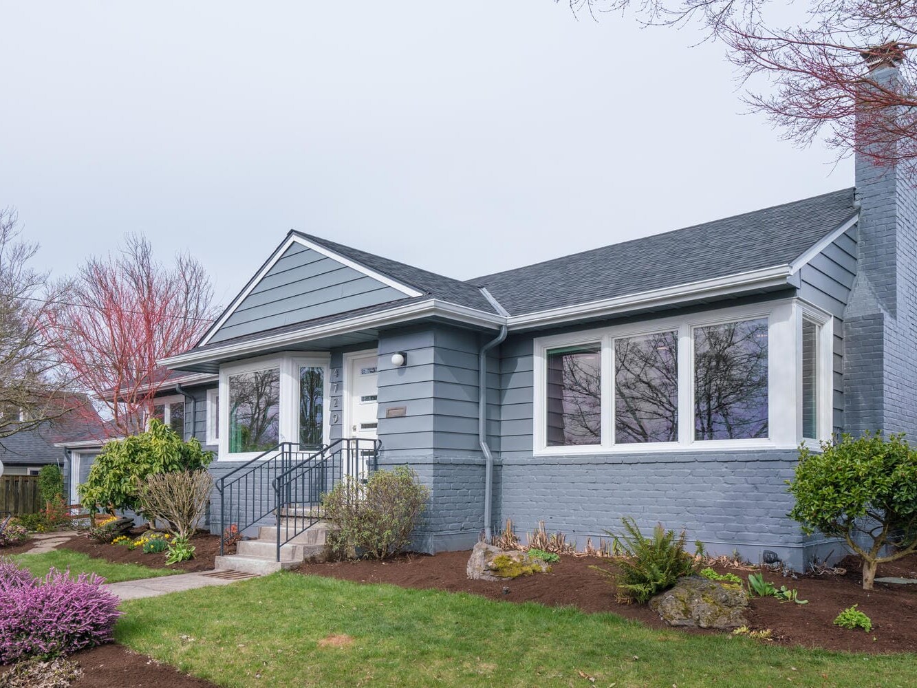 A gray single-story house with a gabled roof, large windows, and a chimney sits quietly in Portland, Oregon. The landscaped yard features trimmed bushes, bare trees, and a pathway leading to the front steps under an overcast sky.