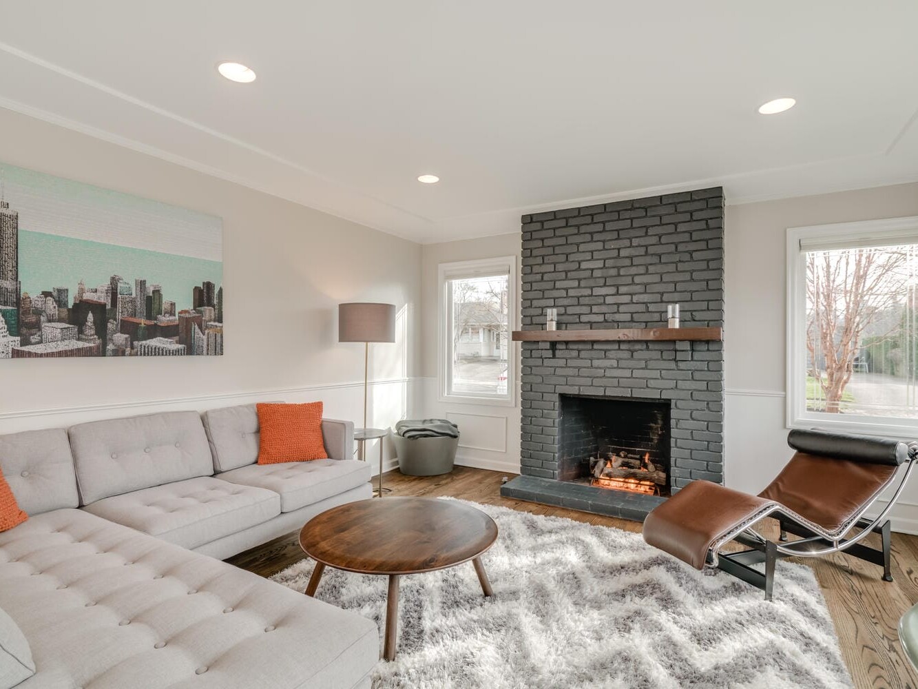 A modern living room in Portland, Oregon, features a gray sectional sofa with orange cushions and a round wooden coffee table on a fluffy rug. A black brick fireplace is in focus, flanked by two windows. A lounge chair and a floor lamp complete the cozy setting.