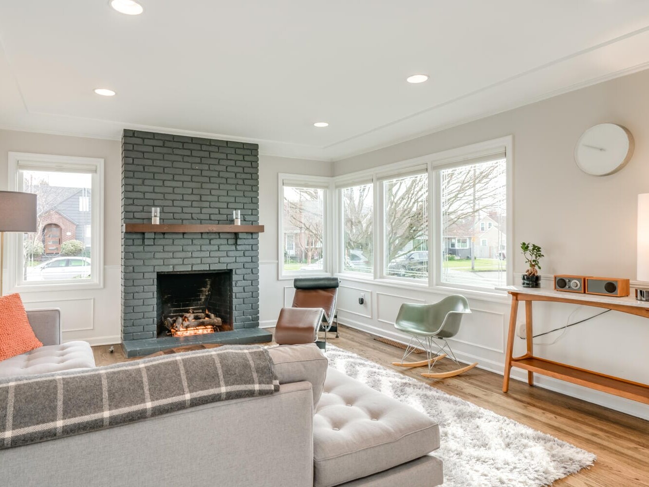 A cozy Portland, Oregon living room features a gray brick fireplace, a gray couch, and a green rocking chair. Large windows fill the space with natural light. A wooden console holds a lamp and record player, while a fluffy rug lies on the wooden floor.