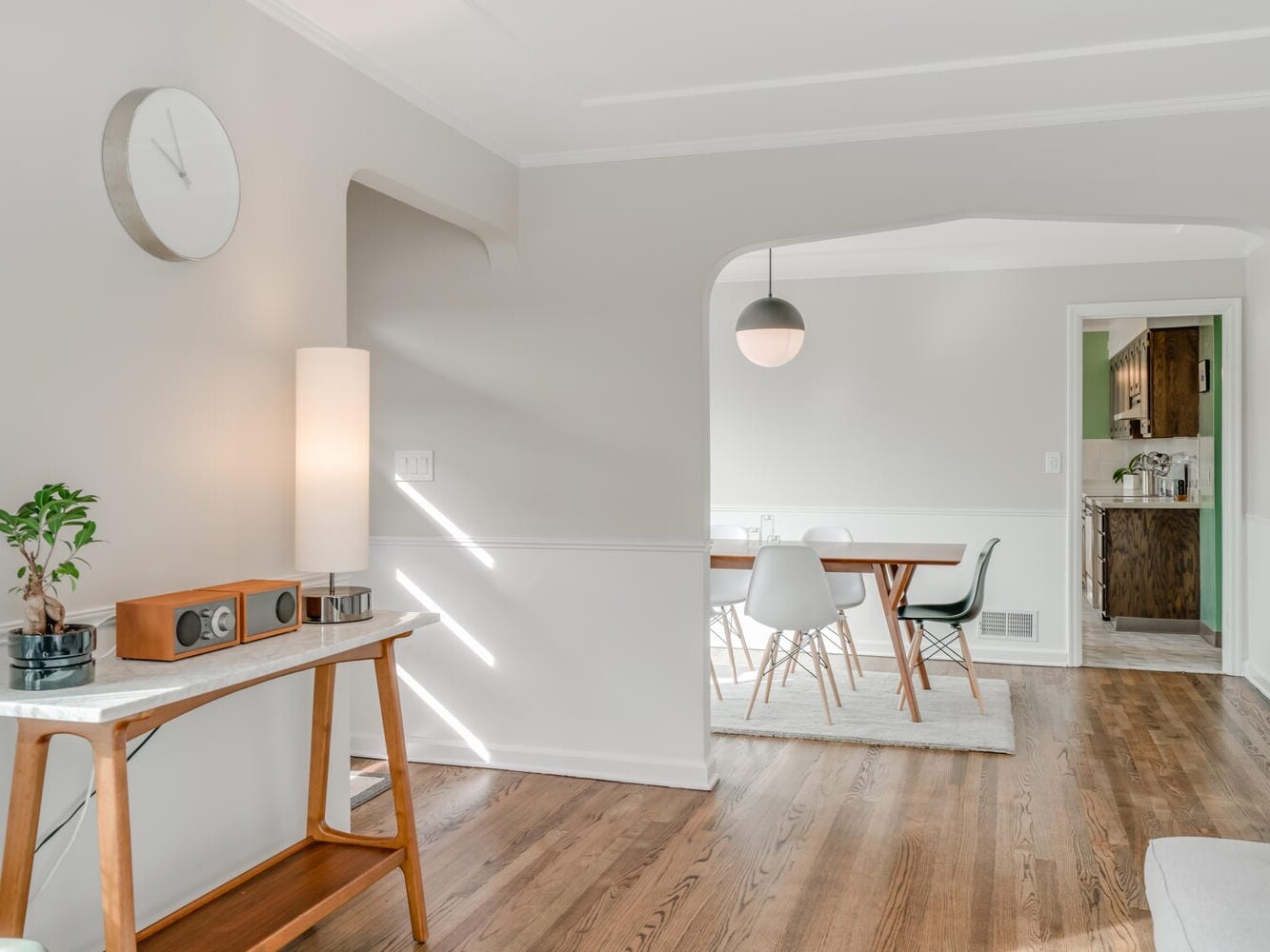 A bright, airy room in Portland, Oregon, with wooden floors features a small table with a plant and speaker, a large clock on a light gray wall, and an archway leading to a dining area with a table, chairs, and pendant light. Natural light streams through the windows.