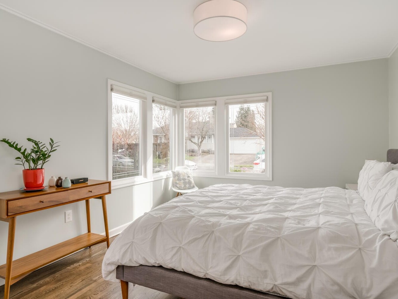 A bright Portland, Oregon bedroom with a large bed featuring a white quilted comforter. The room has light gray walls, a wooden side table with a plant and books, and three windows allowing natural light to fill the space. A modern ceiling light hangs above.
