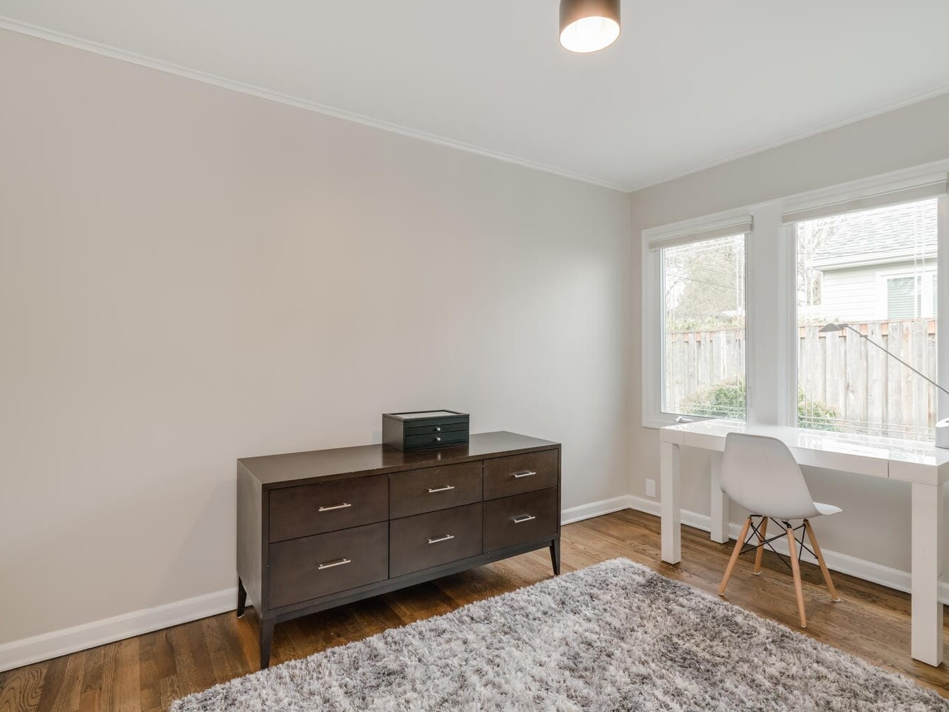 A minimalist room in Portland, Oregon features a wooden dresser, a small printer, and a white desk with a chair beside a window. The light-colored walls complement the gray rug on the wooden floor, and a ceiling light adds brightness to this serene space.