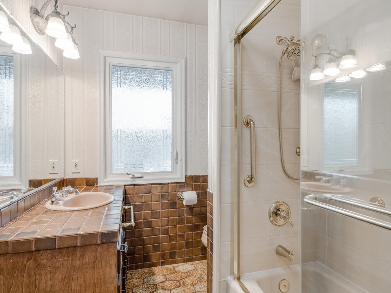 A bathroom in Portland, Oregon, with a wooden vanity and tiled countertop, featuring a sink and mirror. The walls have light wallpaper, and the floor is tiled. A walk-in shower with glass doors sits on the right, while a frosted window lets in natural light.