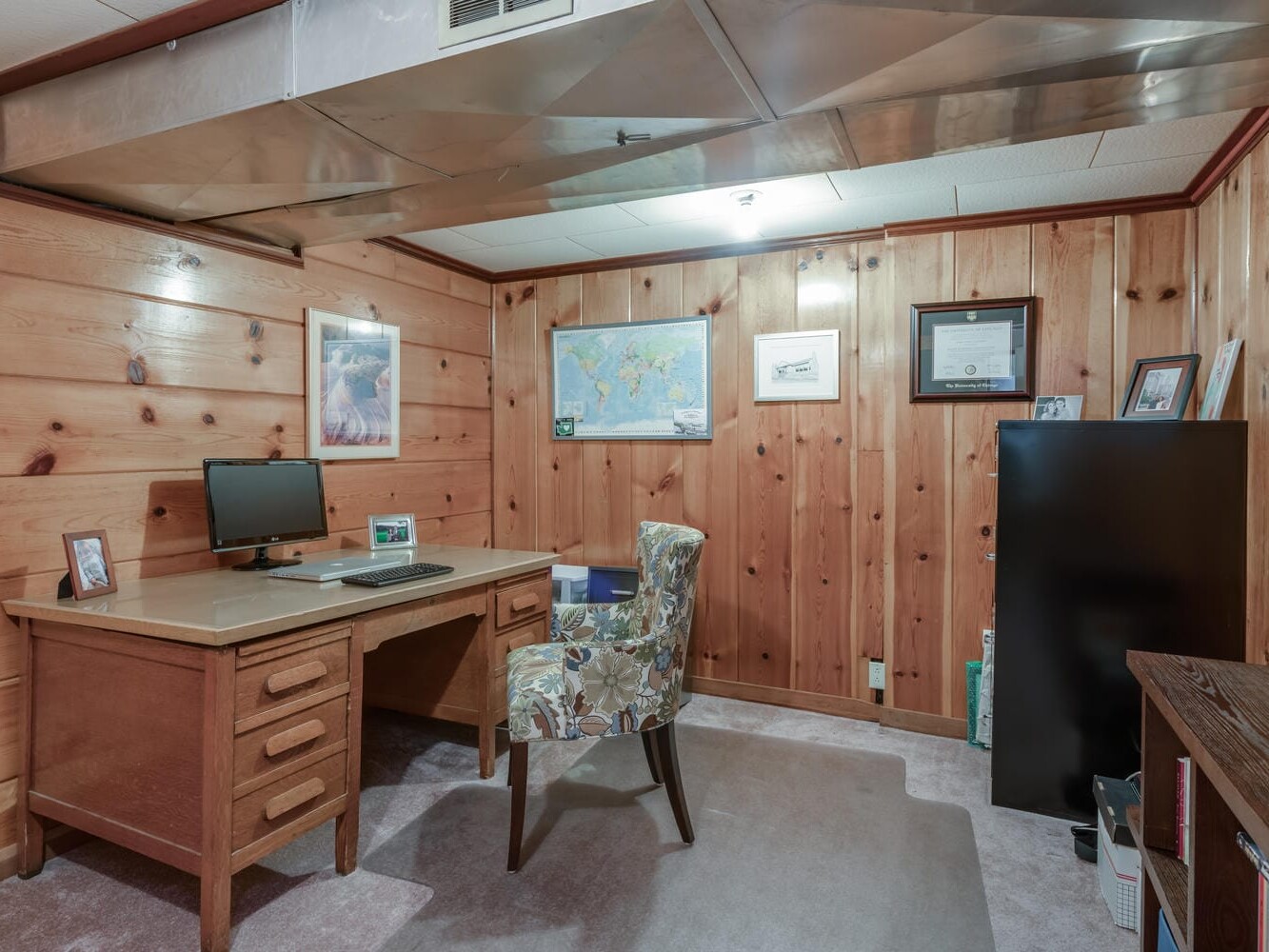 A cozy Portland, Oregon home office with wood-paneled walls features a wooden desk with a computer, floral-patterned chair, and decorative items. Wall decor includes a world map, framed pictures, and a certificate. A file cabinet stands next to the desk.