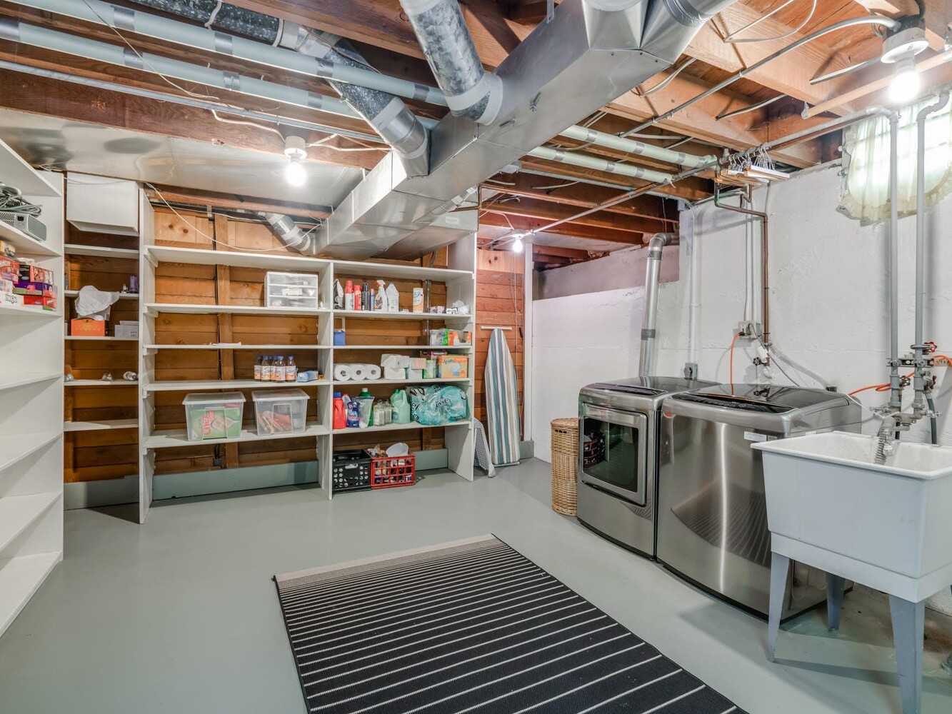 A tidy Portland, Oregon basement laundry room with exposed wooden beams and ducts features a washer and dryer, utility sink, shelves stocked with household items, and an ironing board. A striped rug is placed on the concrete floor.