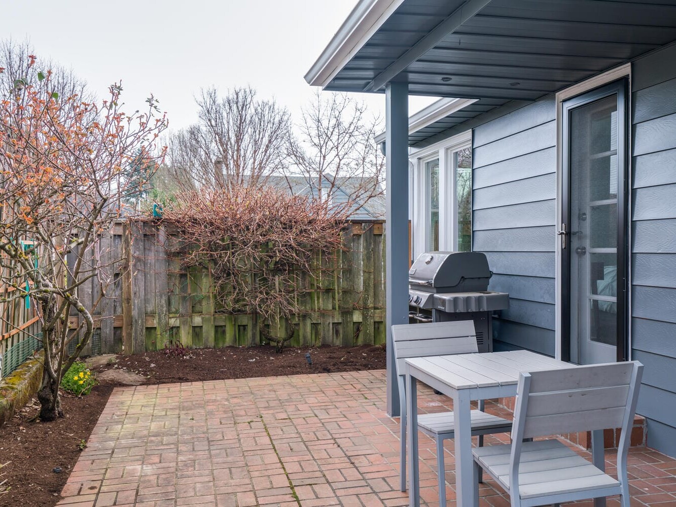 A small backyard patio in Portland, Oregon, features a brick floor, a wooden table with two chairs, and a barbecue grill. The yard is enclosed by a wooden fence, with leafless bushes and trees dotting the perimeter against the light blue house exterior.
