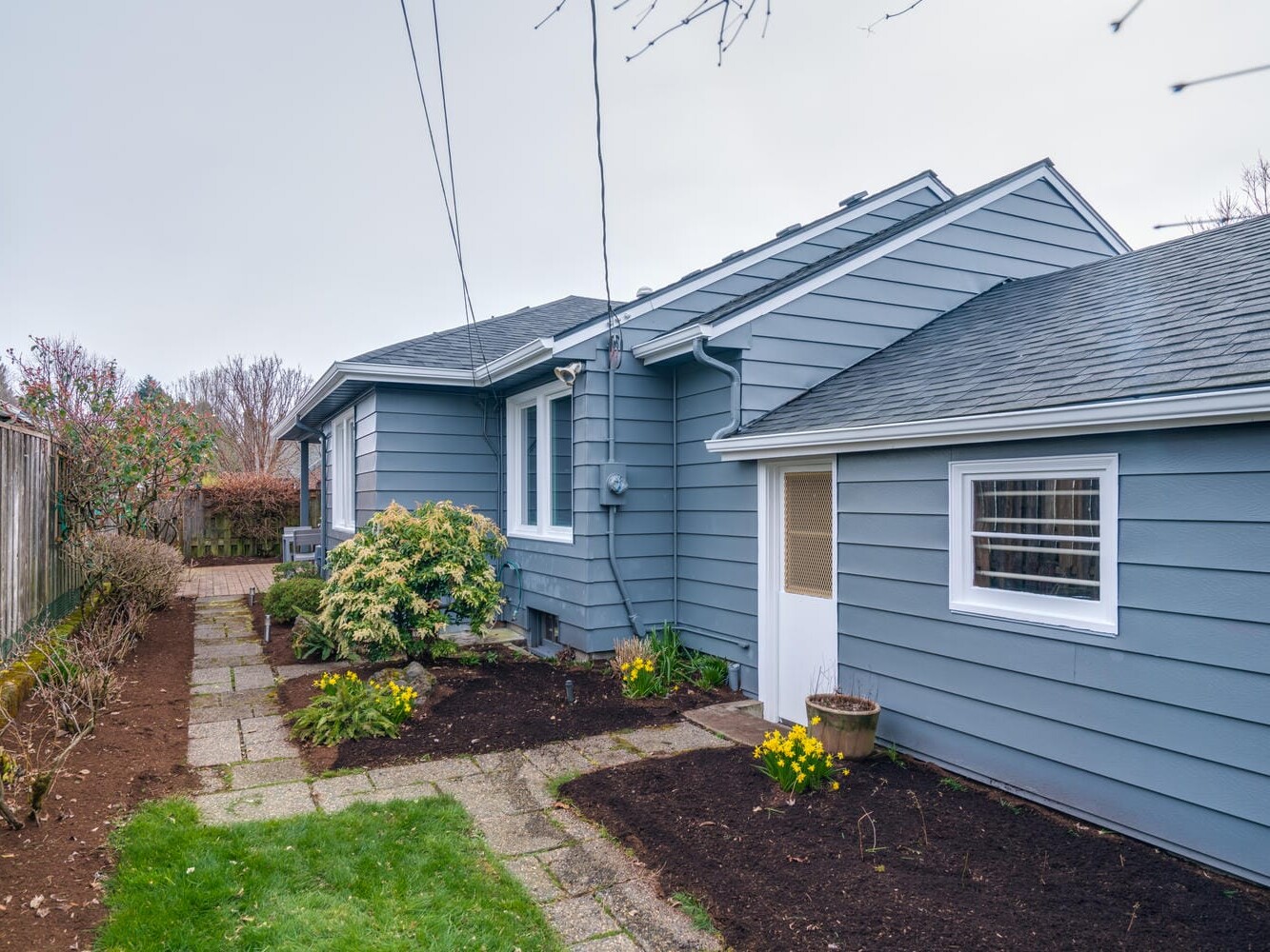 Blue, single-story house in Portland, Oregon with a sloped roof and white-framed windows. A pathway of stone tiles leads to the backyard. The garden features small yellow flowers and green shrubs alongside the house, with a wooden fence to the left.