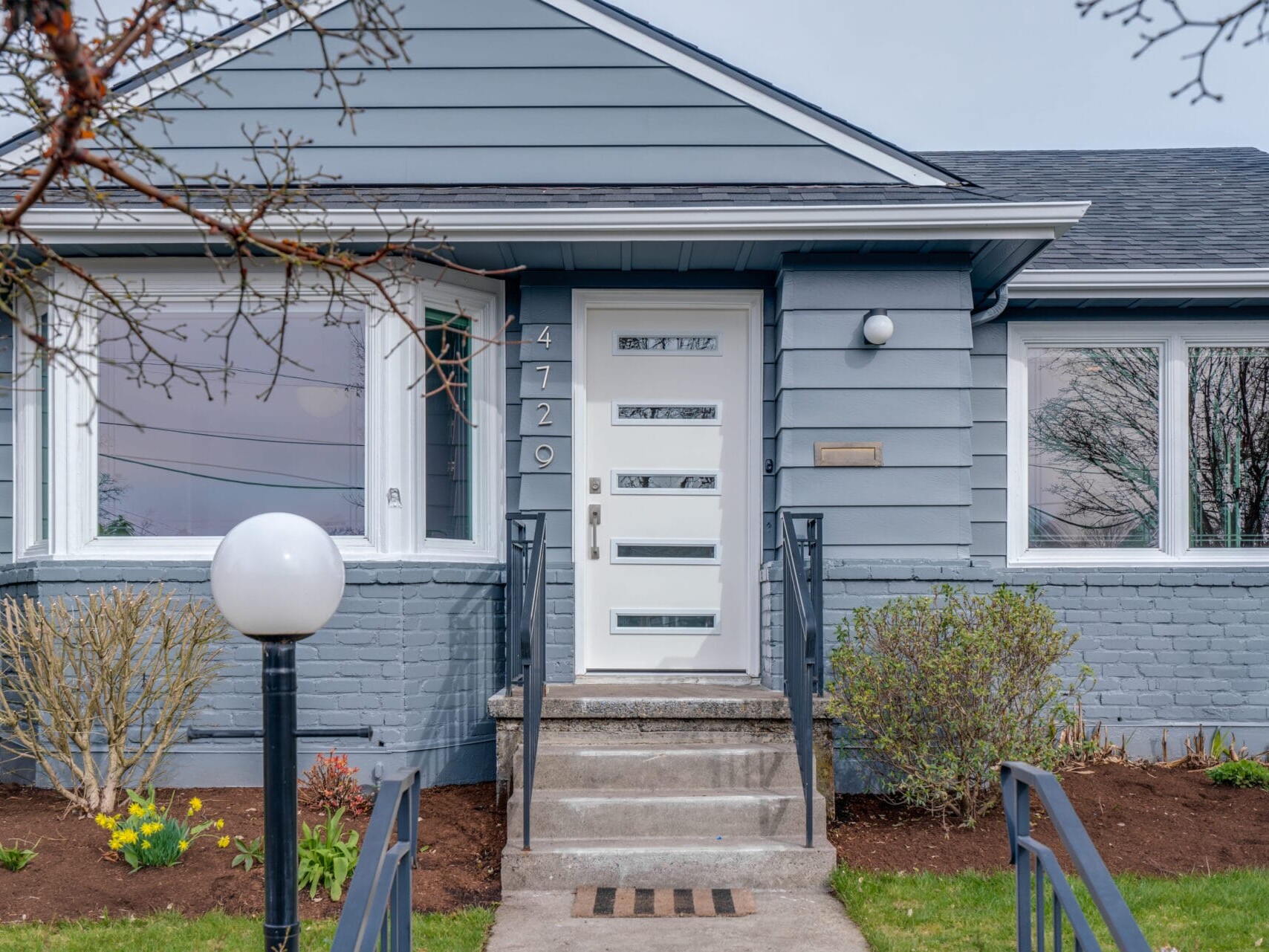 A modern gray brick house in Portland, Oregon features a stylish white front door with decorative glass panels. Two small staircases lead up to the entrance, flanked by a round white lamp post on the left and a beautifully landscaped front yard adorned with flowers and shrubs.