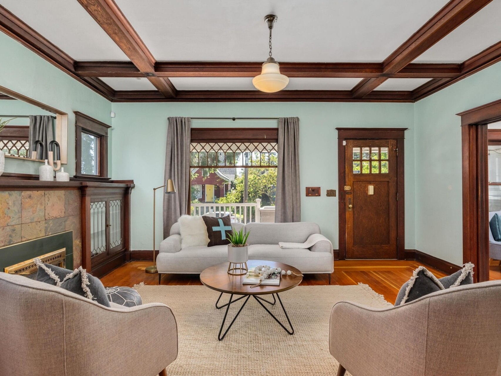 A cozy living room featuring a light blue wall, wooden ceiling beams, and a brown door. There is a gray sofa with a black and white pillow, two beige armchairs, a round coffee table, and a fireplace with decorative items. A window lets in natural light.