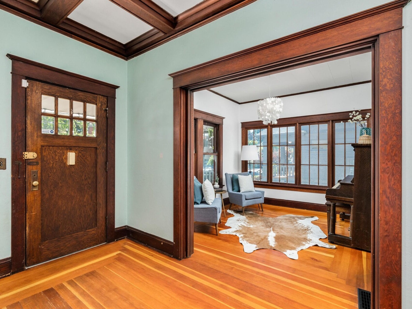 A cozy room with hardwood floors, a wooden front door, and a seating area featuring blue chairs. A cowhide rug lies on the floor, and a piano is near the large windows letting in natural light. A white pendant light hangs from the ceiling.