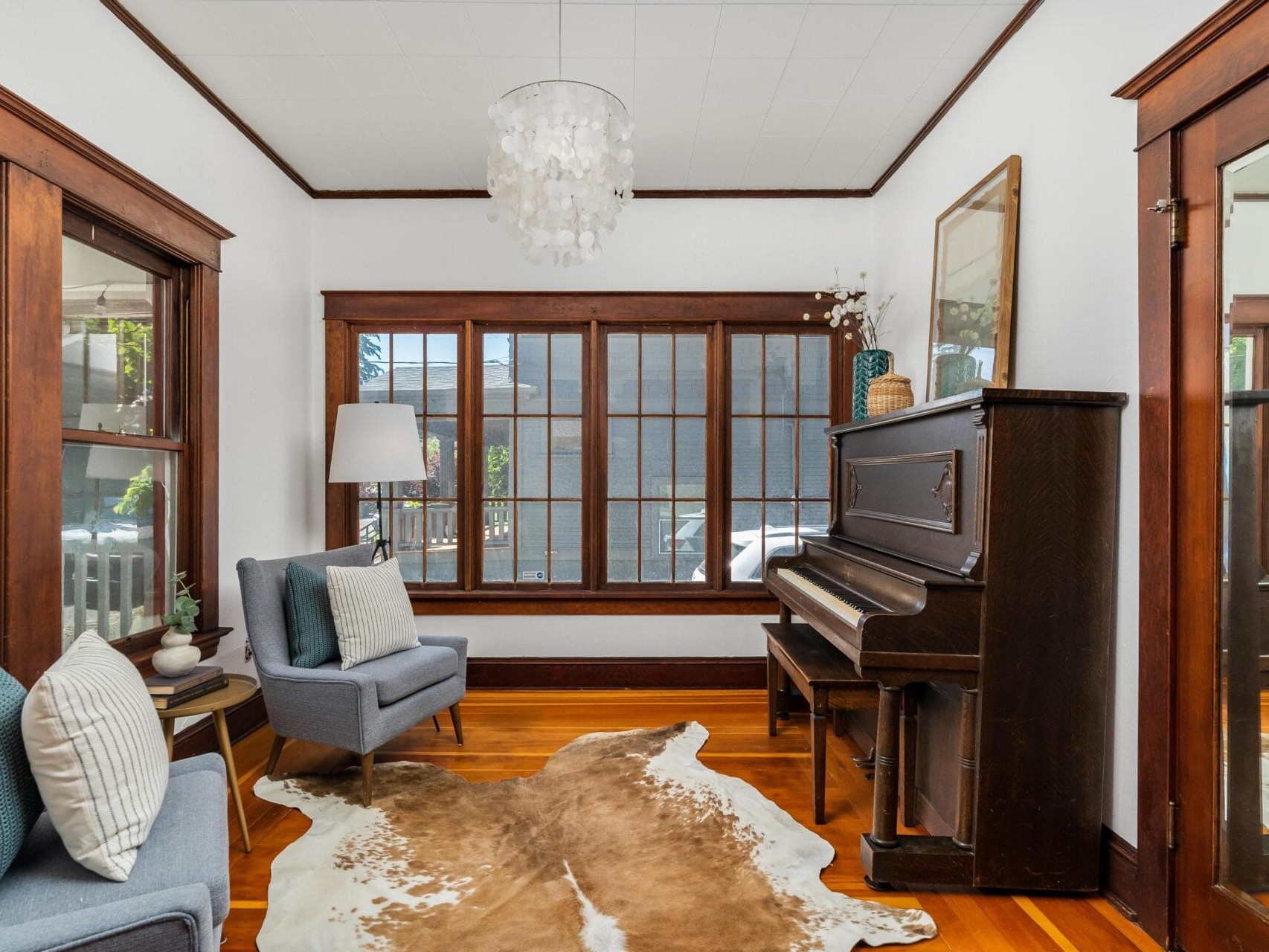 A cozy living room with a dark wooden piano on the right, two gray upholstered chairs with cushions on the left, and a cowhide rug on a wooden floor. Large windows line the back wall, and a modern chandelier hangs from the ceiling.