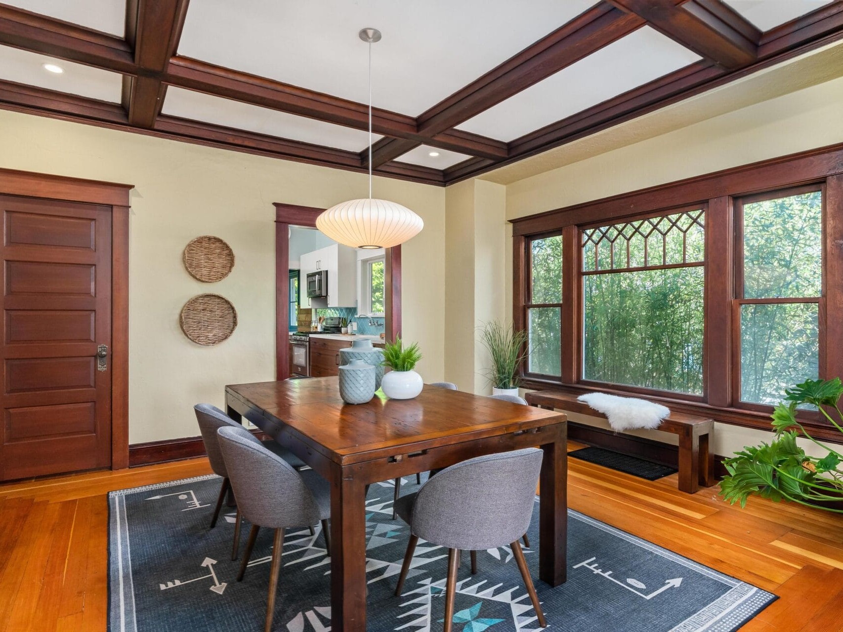 A dining room with a wooden table and four gray chairs. The room has a coffered ceiling, a geometric patterned rug, a pendant light, and large windows with wooden frames. Green plants and wall decor add a touch of nature.