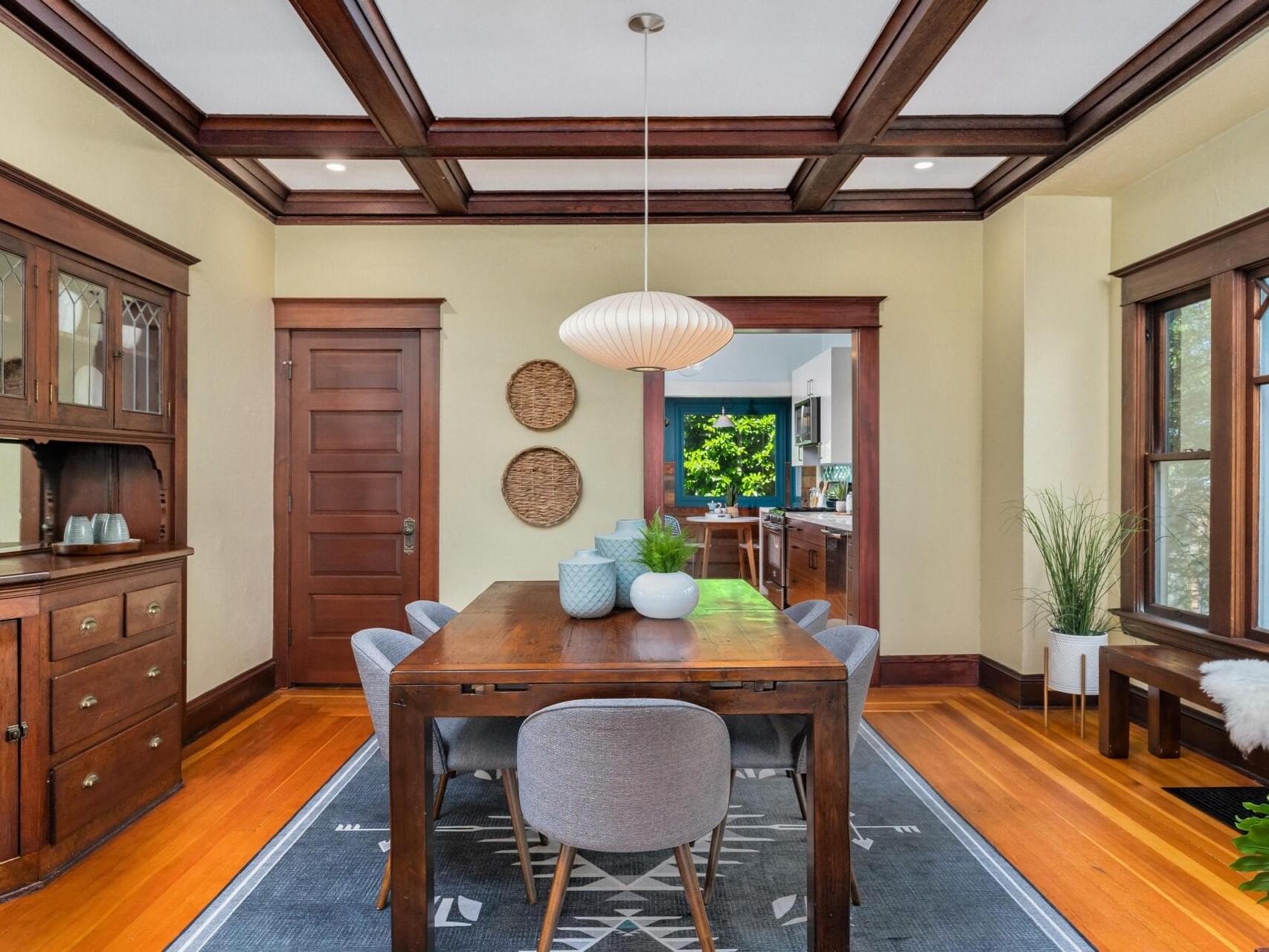 A dining room with a wooden table and six gray chairs. A modern pendant lamp hangs above. The room has wood-paneled walls, a sideboard, two woven wall decorations, a large window, and a view into the kitchen. A blue rug covers the floor.