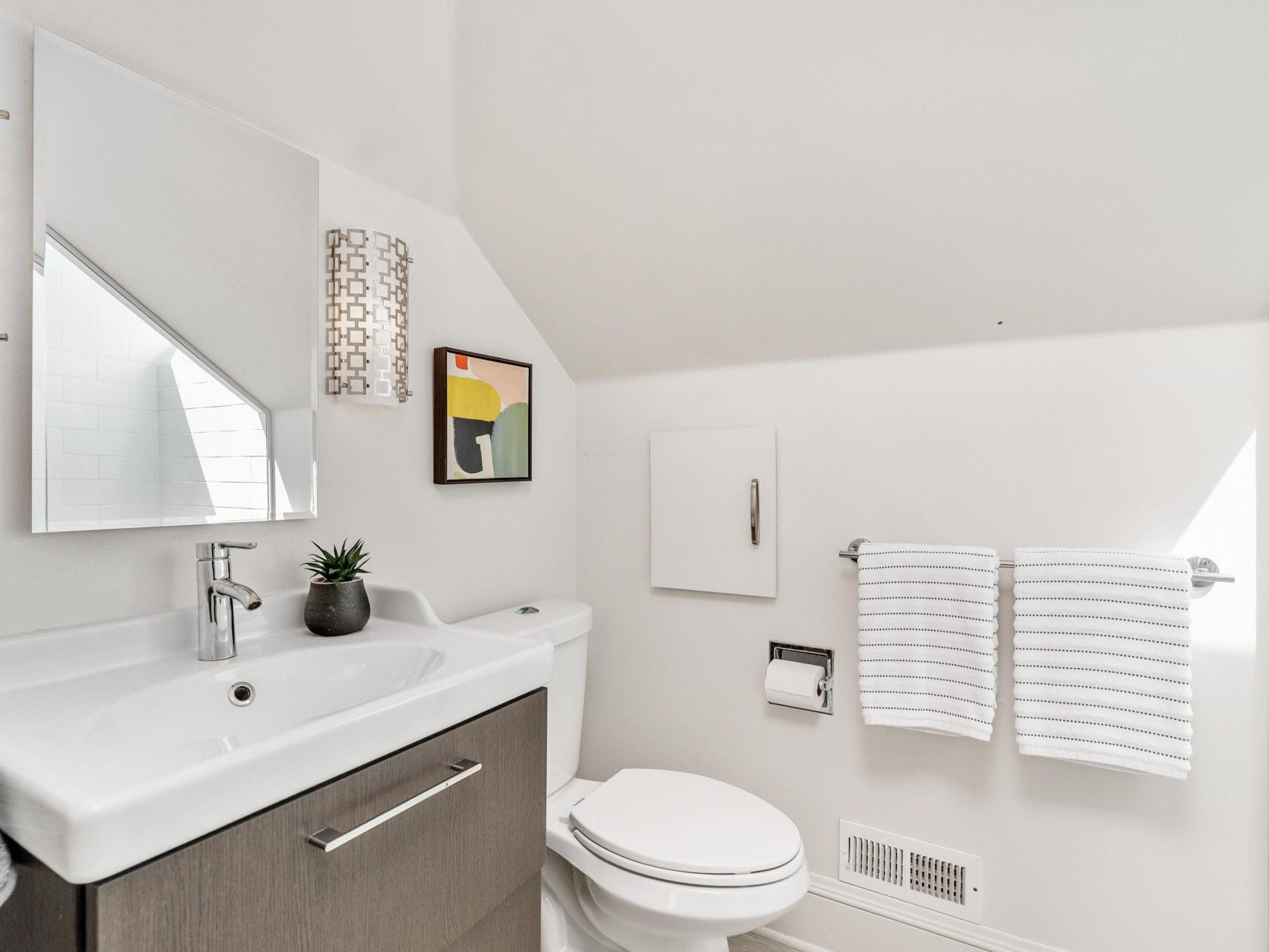 A modern bathroom with a white sink and a faucet on a dark wood vanity, a toilet, and a wall-mounted towel rack with two striped towels. Theres a small potted plant on the sink and a colorful abstract painting on the wall.