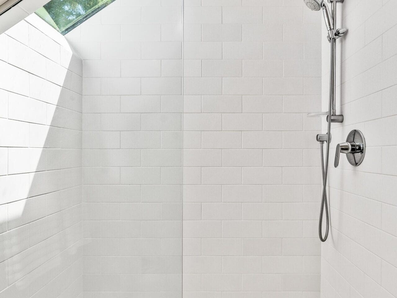A modern, minimalist shower features white tile walls and a glass door. It includes a sleek showerhead and handle on the right, with natural light streaming in from a skylight on the left. The design is clean and bright.